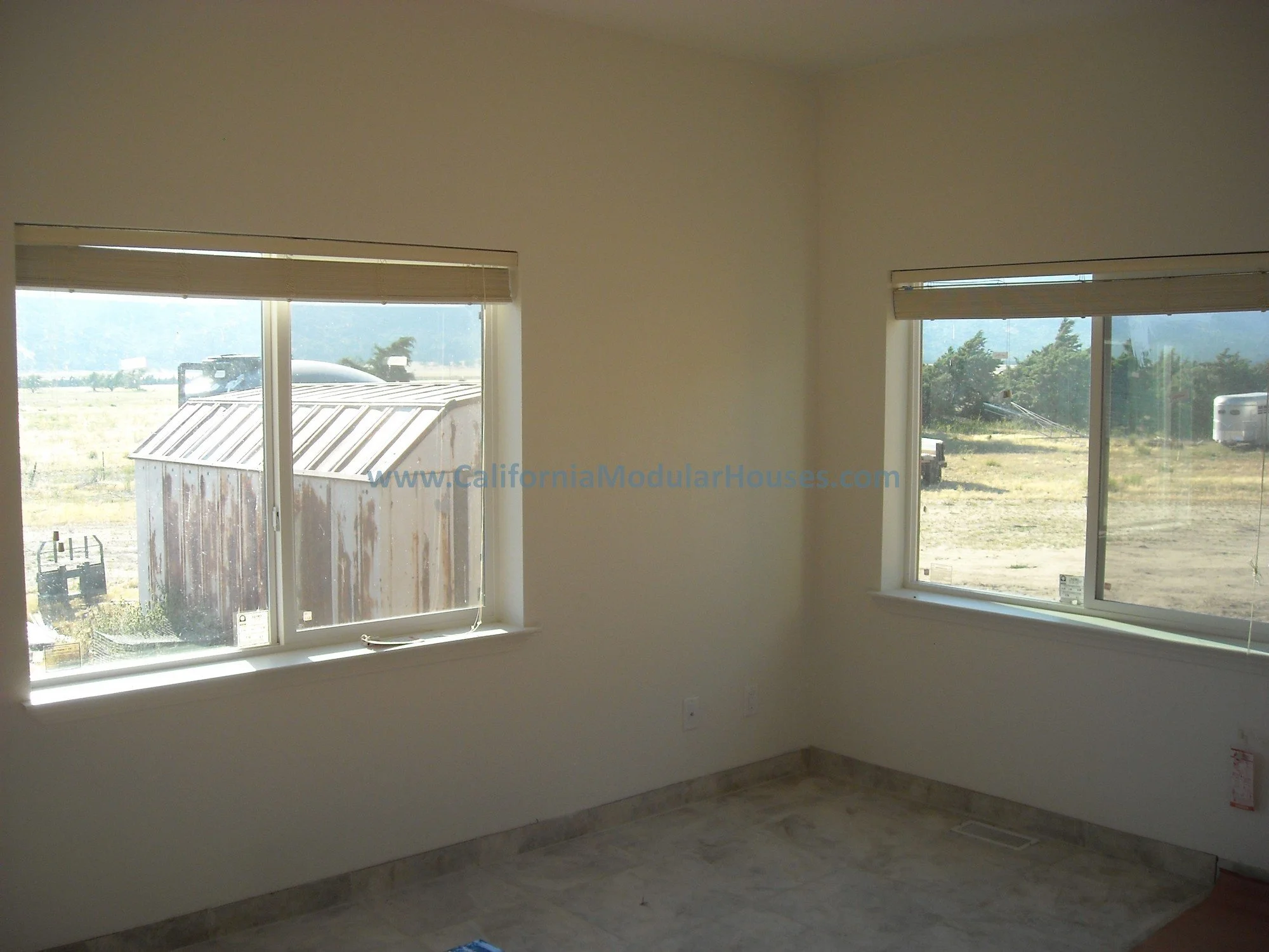 Bedroom with two large windows showing an outdoor landscape with a rusted shed and open field showing the cattle ranch.  Factory Built Housing for California.   