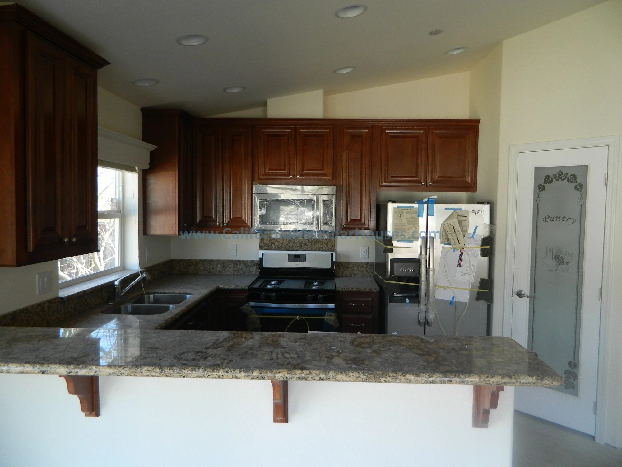 Kitchen with wooden cabinets, granite countertops, a window over the sink, a stove, microwave, and a refrigerator taped for renovation, with a pantry door on the right.