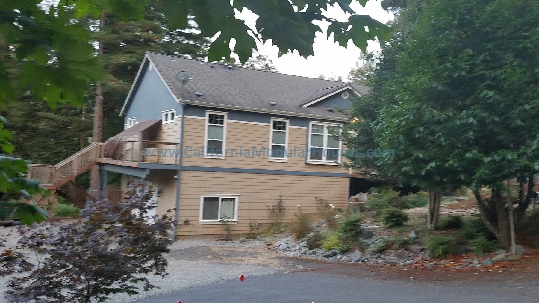 A two-story house with gray and beige siding, multiple windows, surrounded by trees and plants, with a gravel driveway. Del Norte County Modular, Modular Homes California,
