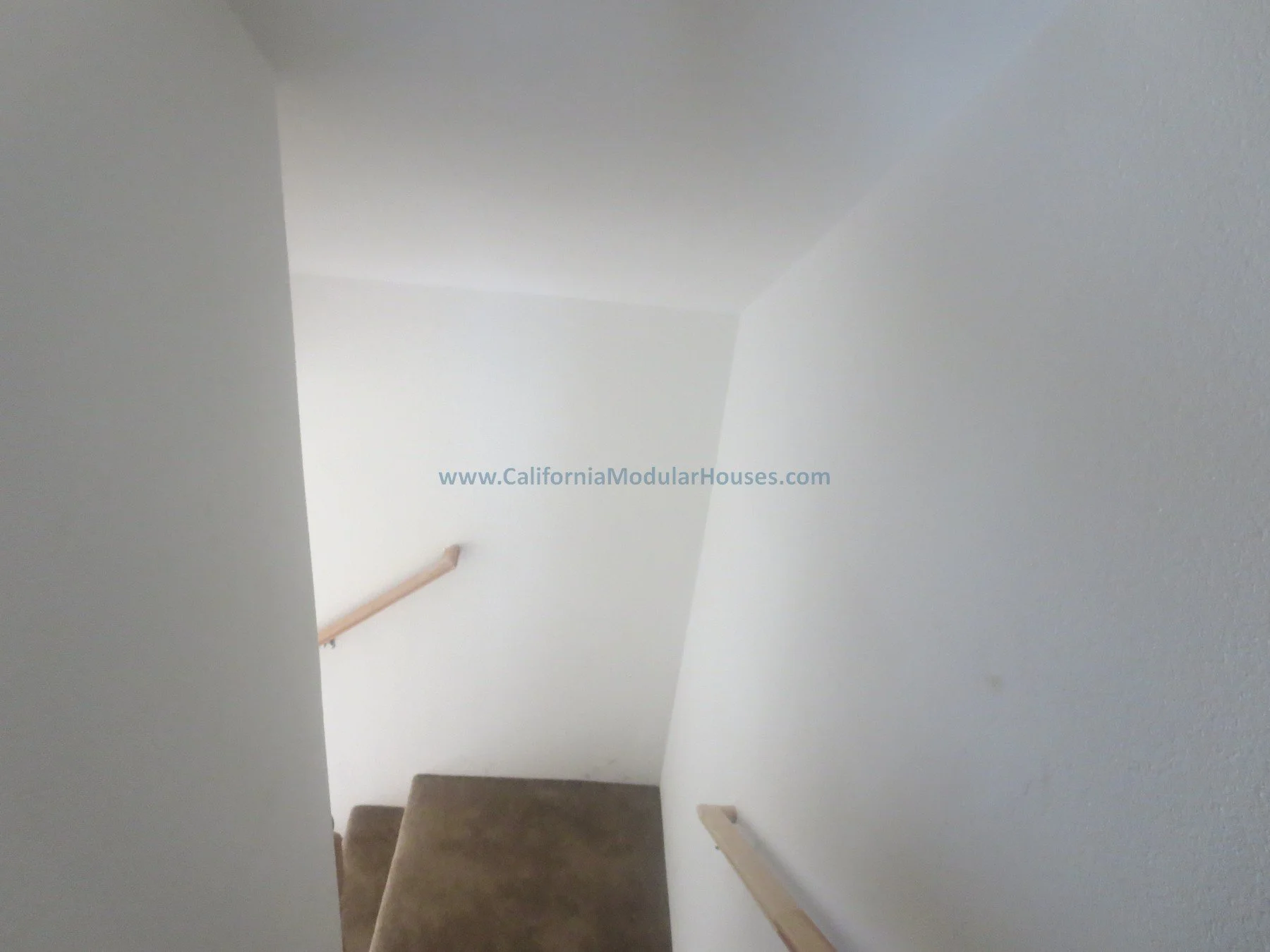 Unfinished attic.  New pre-fab modular house in Napa, California.  California modular home.  A stairway with brown carpeted steps and wooden handrails on both sides, viewed from the top looking down, with white walls. www.CaliforniaModularHouses.com.