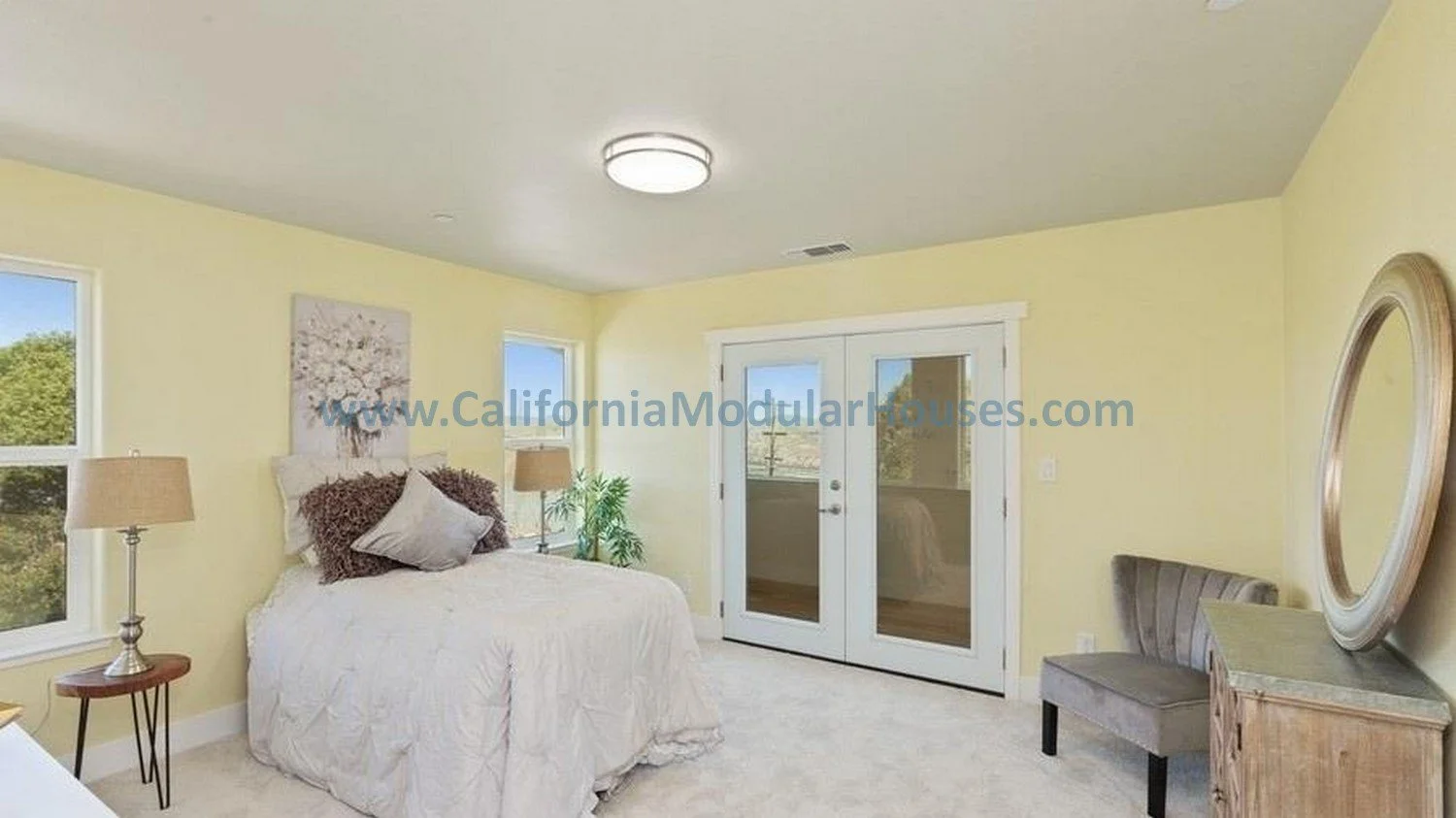 Bright bedroom with yellow walls, white bed, and French doors leading outside, with two windows, lamps, a plant, a bench, and a mirror.  California Modular Houses, Inc.  Modular house interior.  
