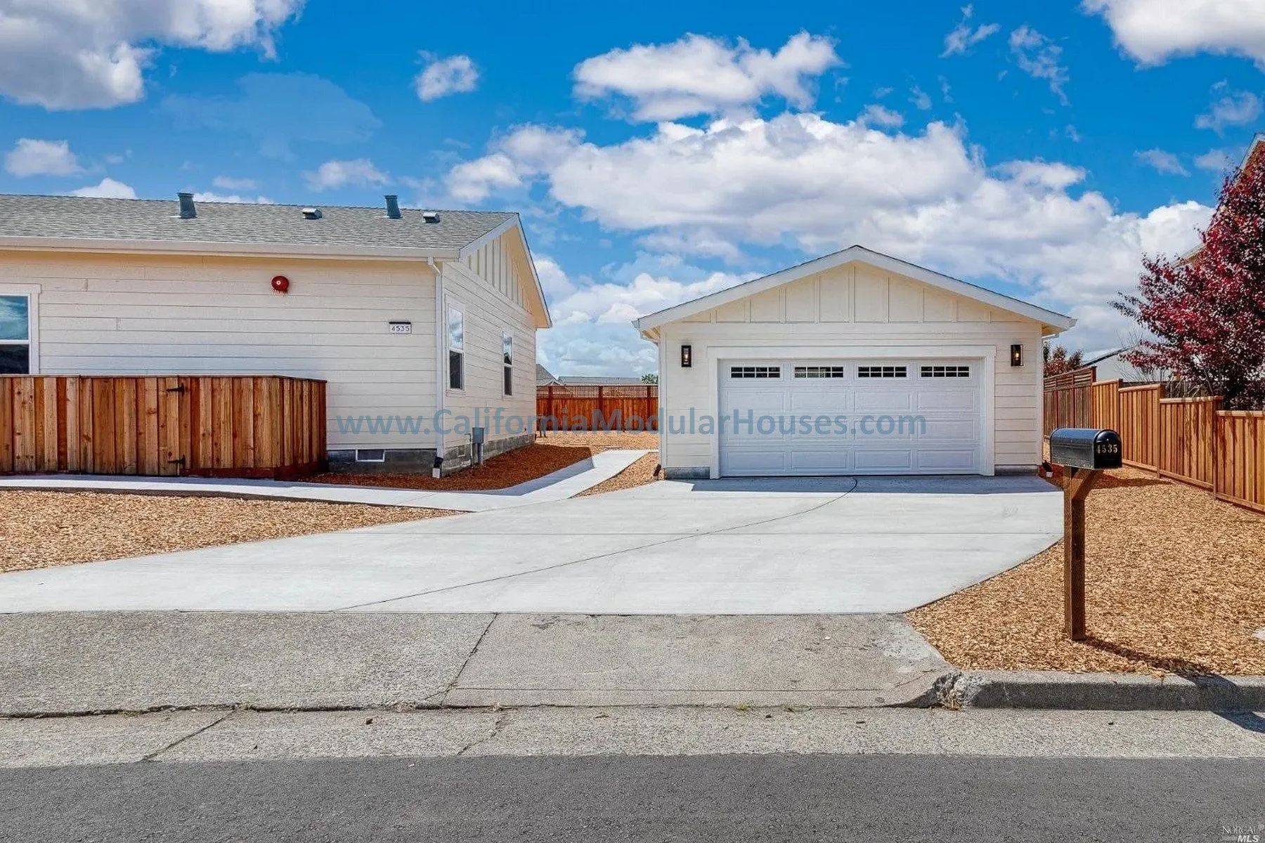A modern house with a white garage, fenced yard, and a mailbox in front, under a blue sky with clouds.
