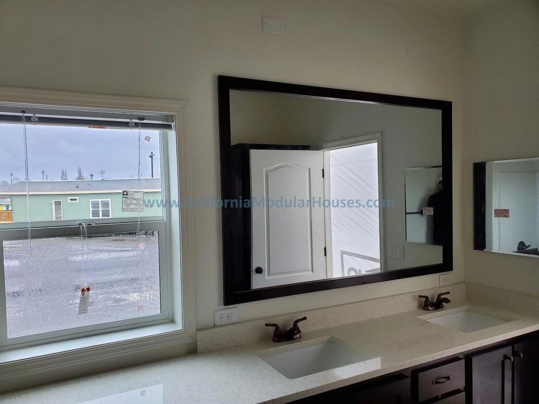 Bathroom with double vanity sink, two mirrors, and window showing a rainy day outside. Modular Homes in California, Prefab Home California, Pre-Fabricated Homes,