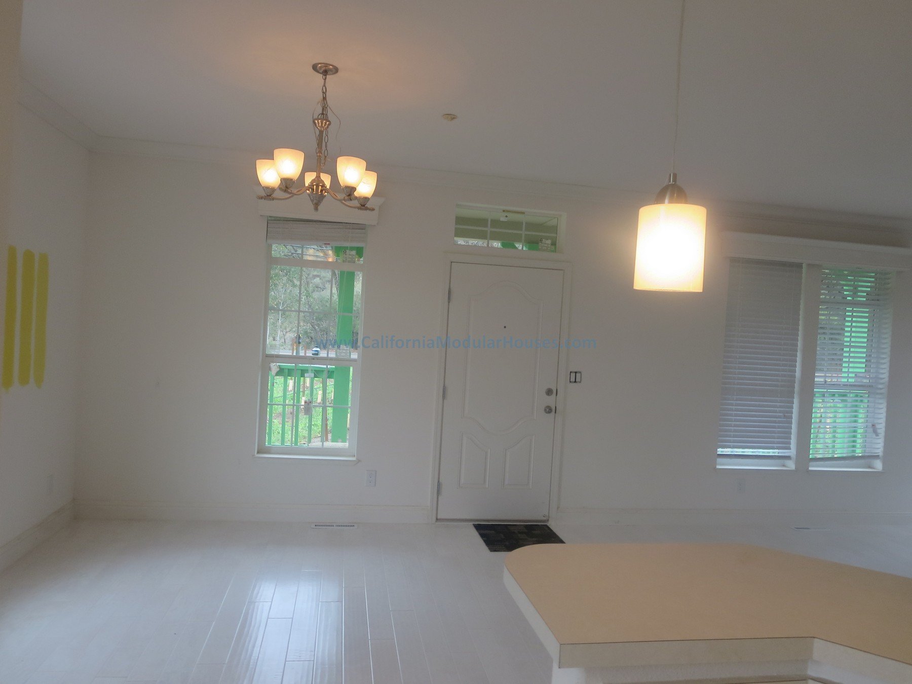 Empty living room with white walls, two windows with blinds, a chandelier, a ceiling light, a front door, and a small part of a kitchen counter visible.  Modulars.   Napa, CA.  Prefab.  California Modular House. California modular home.  Modulars.  