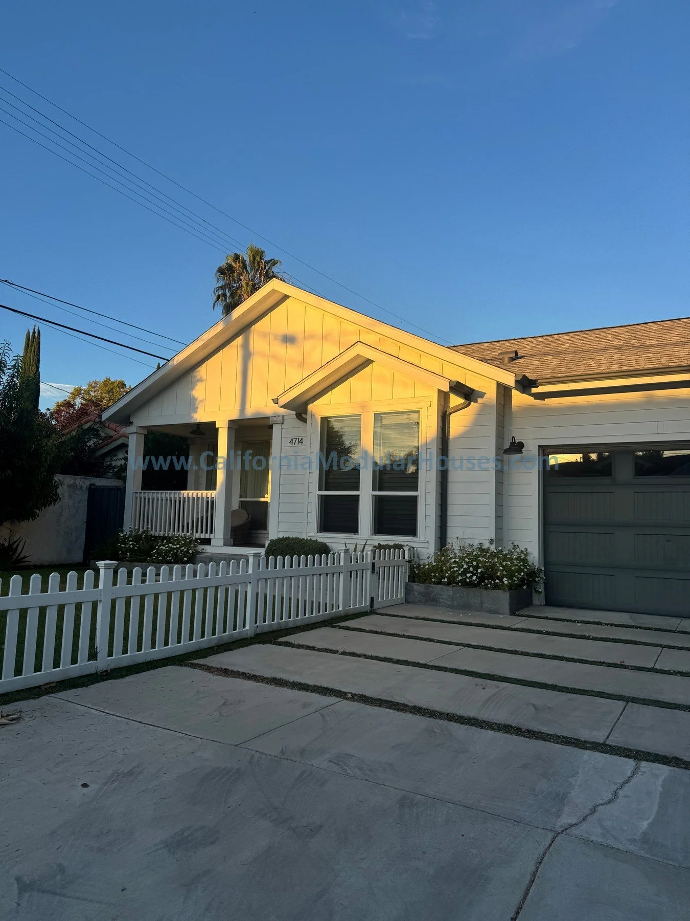 White modern house with a front porch, a white fence, and a driveway with concrete slabs and grass strips, under a clear blue sky.