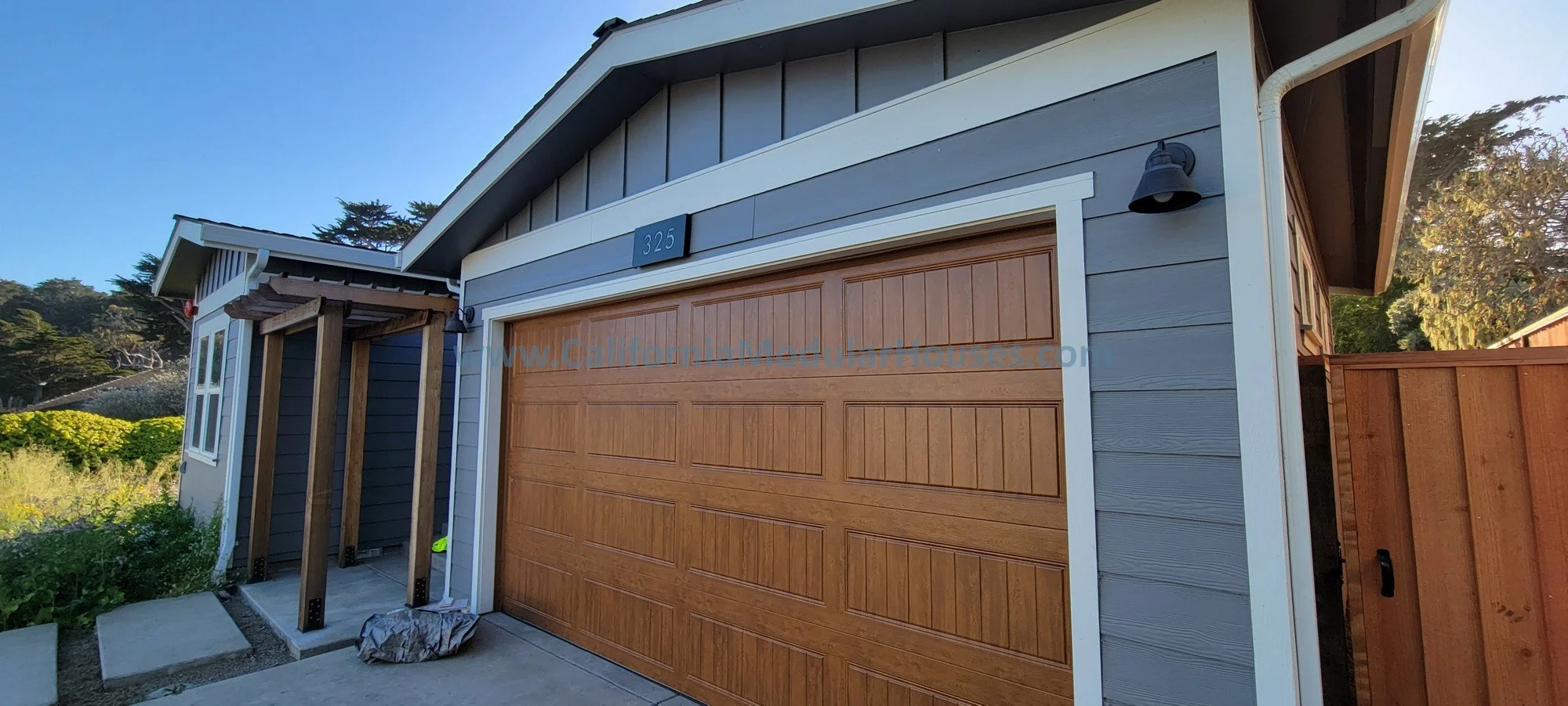 Newly constructed house with grey siding, a wooden garage door, and a small entrance porch with wooden beams, set in a natural outdoor environment.