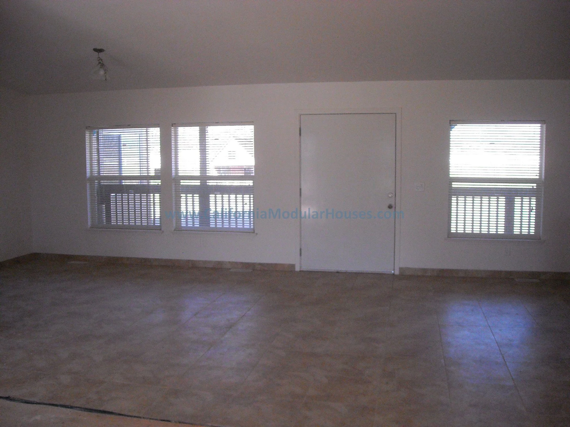 Empty room with three windows and a door, beige tiled floor, and white walls.