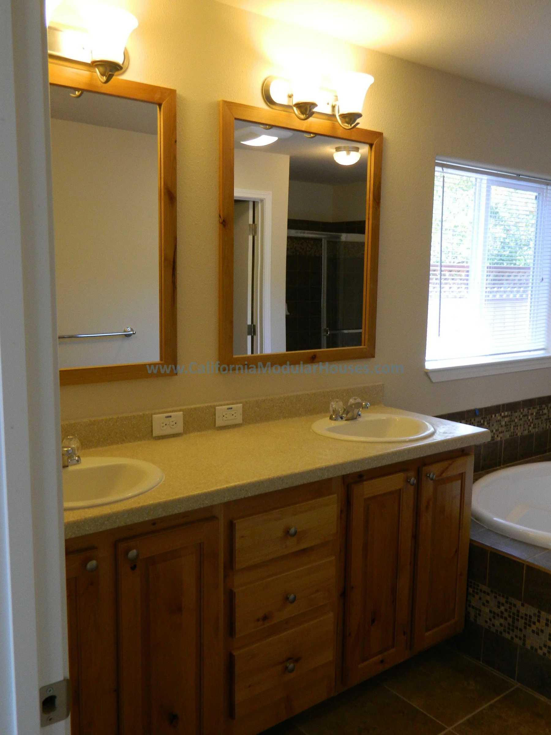 Bathroom with double sinks, wooden cabinets, mirrors, a window, and a tiled bathtub area.