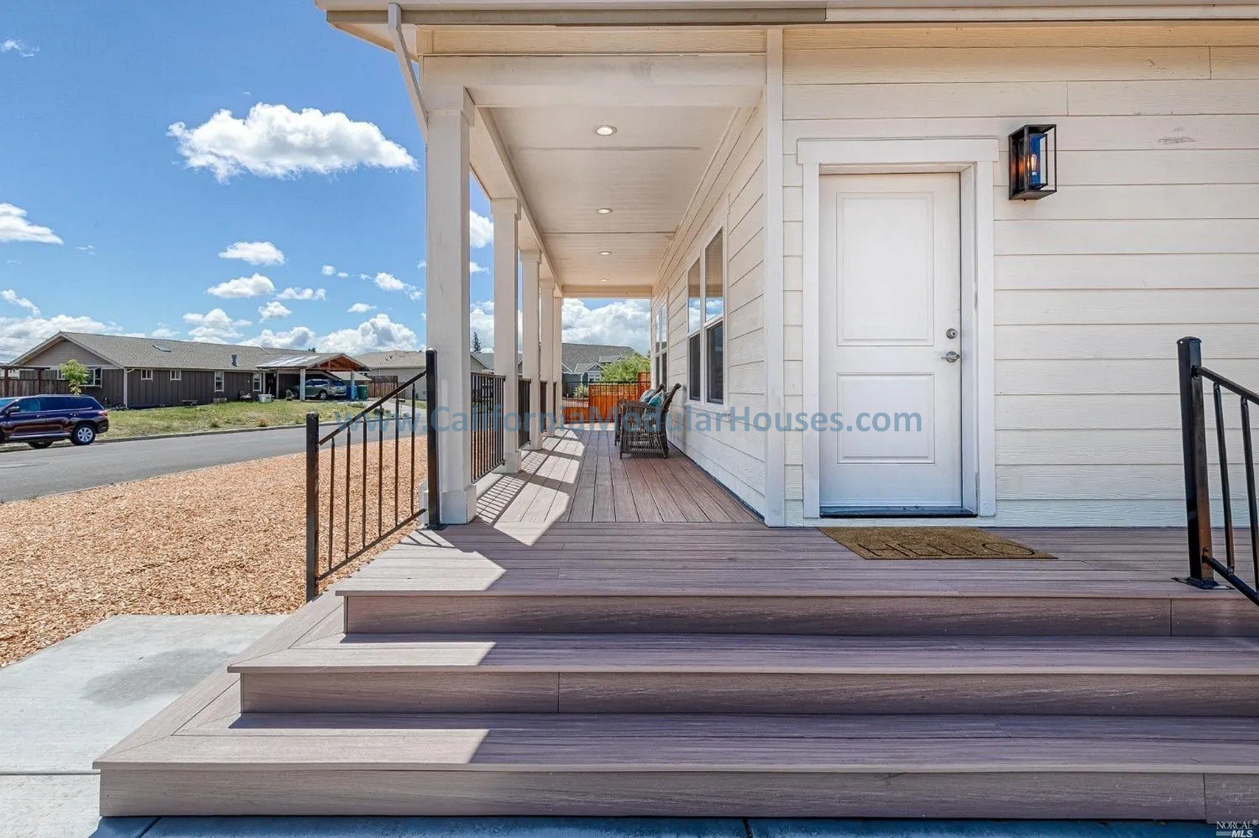 View of a modern house front porch with wooden steps, black metal railing, and white exterior walls, under a blue sky with scattered clouds.