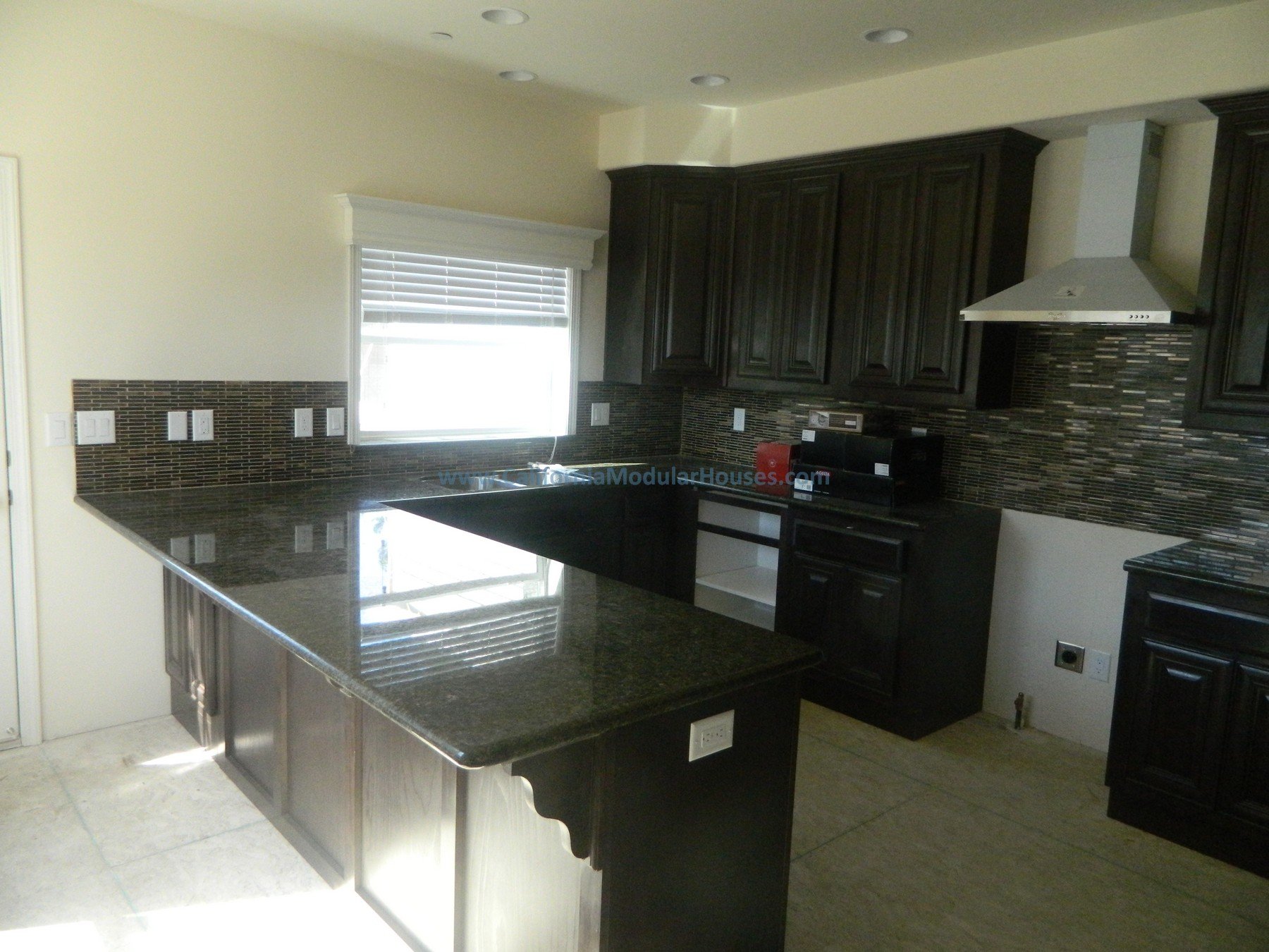 Kitchen with dark cabinets, a black granite countertop island, a window with white blinds, a modern tile backsplash, a range hood, and appliances on the counter.  Prefab Home California, Pre-Fabricated Homes, Modular Homes Bay Area.