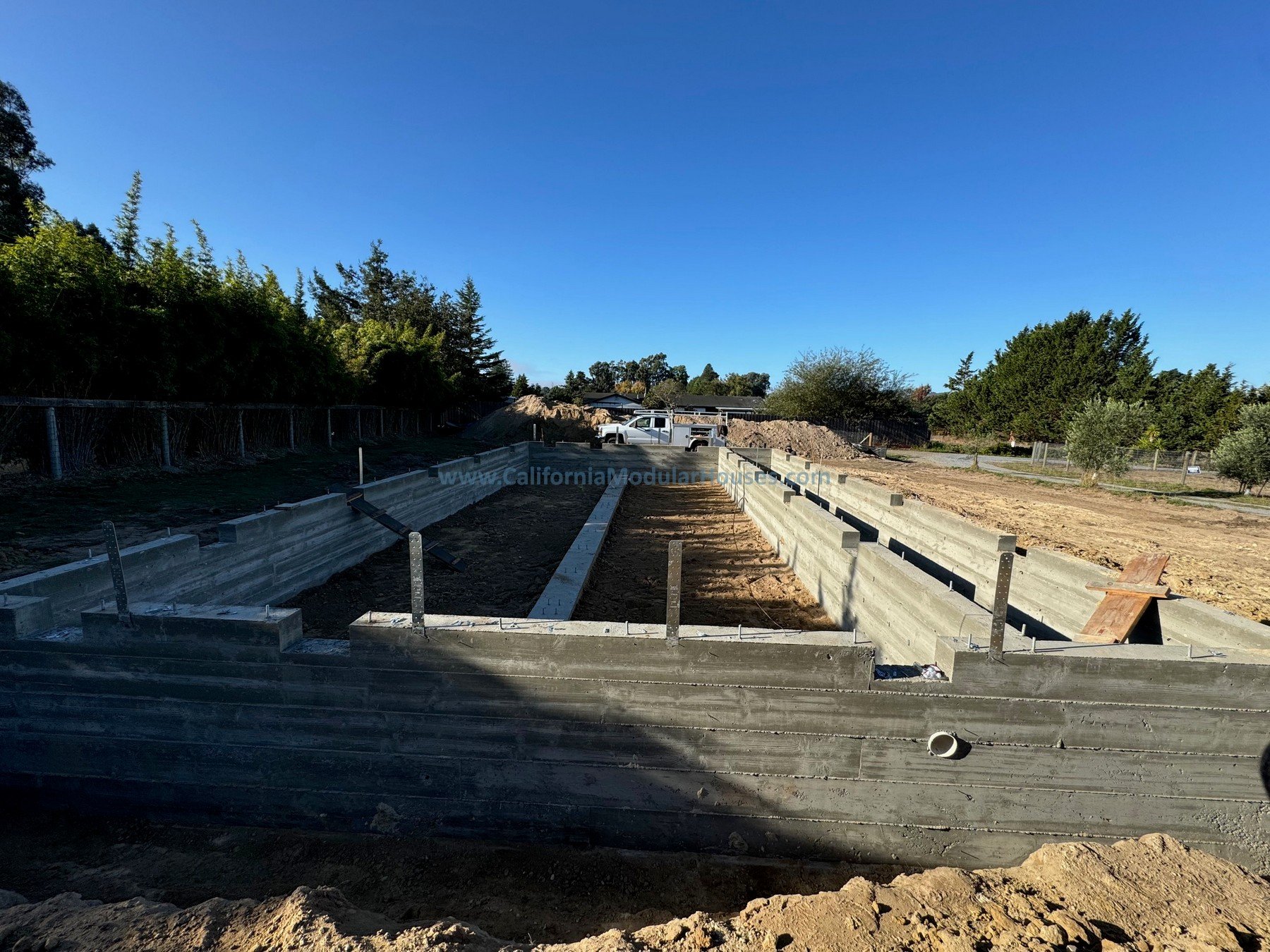 Construction site with concrete formwork in the foreground, and trees and a clear blue sky in the background. Sebastopol, Sonoma County, CA.  Prefab modular Accessory Dwelling Unit foundation.  