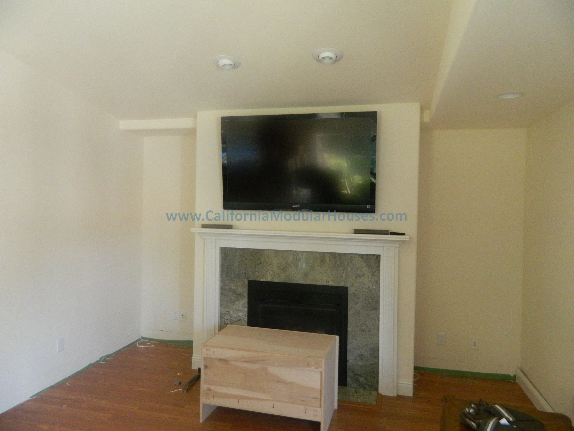 Living room with a mounted flat-screen TV above a fireplace with a green marble surround, a small unfinished wooden table in front, and two smoke detectors on the ceiling.