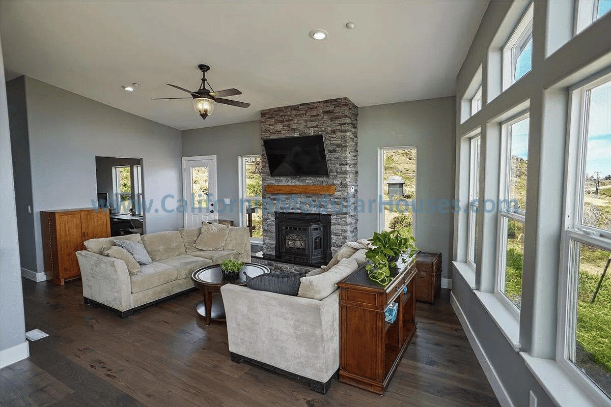 Living room with beige sofas, a stone fireplace with a mounted TV, wooden furniture, and large windows showing an outdoor landscape.