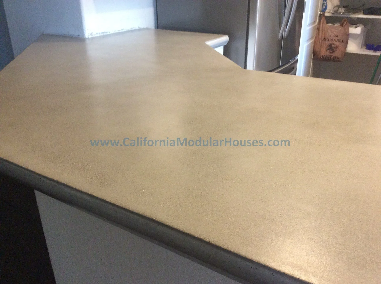 Photo of a beige modular countertop with a website URL printed on it, in a kitchen area with a refrigerator and shelves in the background.