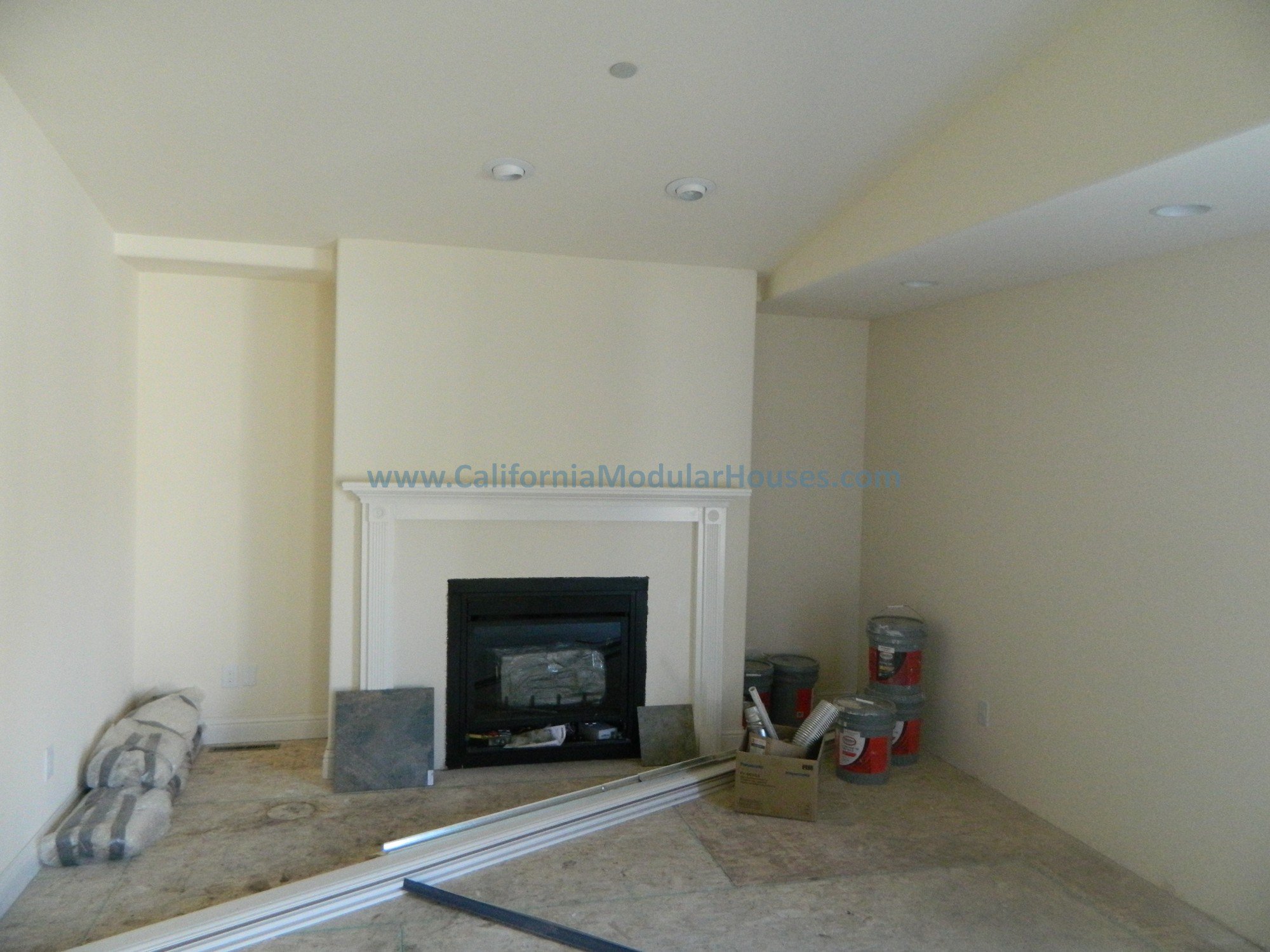 Living room under construction with a white fireplace, construction materials, and recessed ceiling lights.