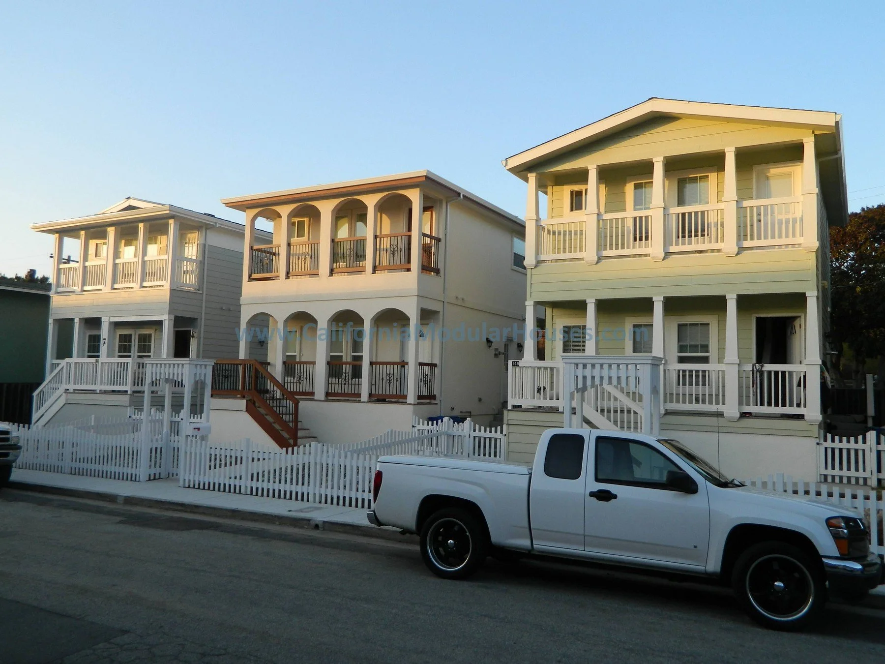 Three multi-story houses with porches and railings, a white picket fence, and a white pickup truck parked in front on the street during daytime.