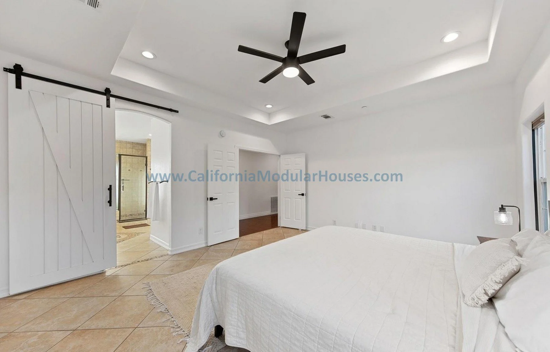 Bright bedroom with white walls, a ceiling fan, and a bed covered in white linens. A sliding barn door leads to a bathroom with a shower. Recessed lighting and a modern lamp are visible.
