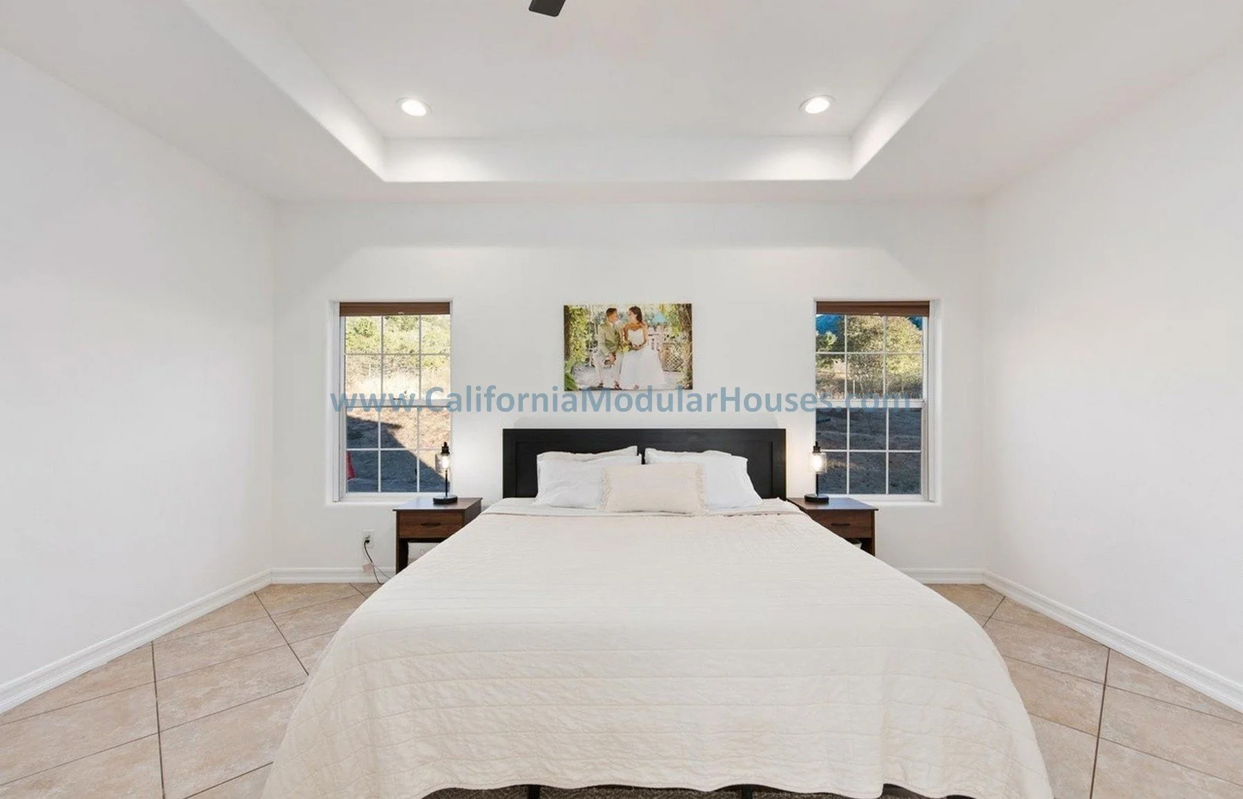 Newly built bedroom with white walls, a small bed with white quilt, two small nightstands with lamps, a framed photograph above the bed, and two windows showing outdoor greenery.
