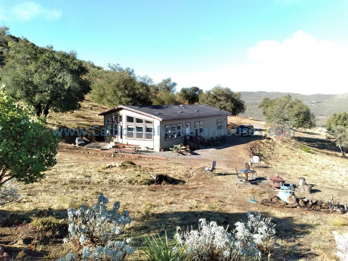 A house on a hillside with trees around, outdoor chairs, tables, and fire pits in the yard, with mountains and a blue sky in the background.