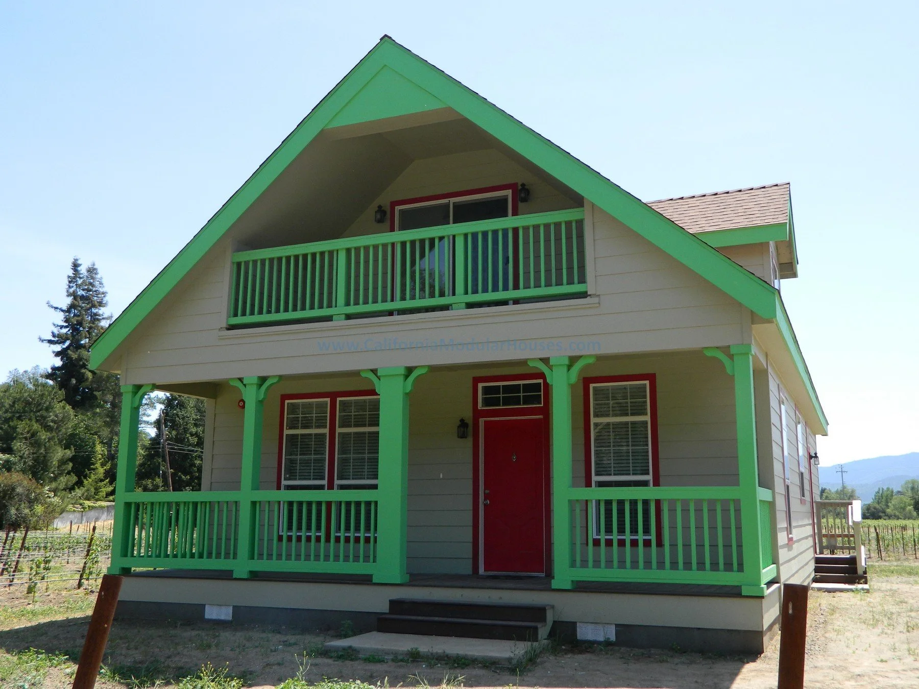 A two-story house with a green trim, red front door, and a balcony, situated in a rural area with trees and mountains in the background.