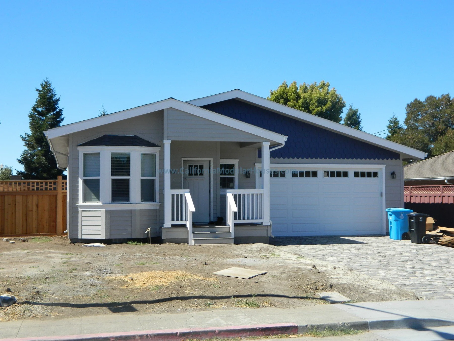 Newly built single-story house with a gray exterior, white trim, and a white garage door, with a small front porch and steps, on a partly fenced lot with a paved driveway.  San Francisco Bay Area, CA.  City of San Mateo, San Mateo County, CA. Modular