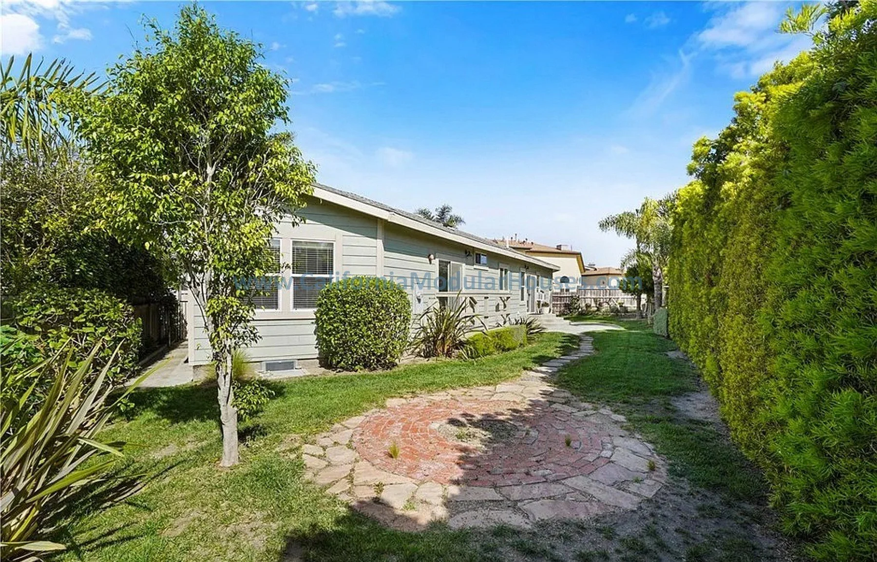 View of a backyard with a green house, lush green trees and bushes, a stone pathway, and a green hedge on a sunny day.  California Modular Houses. Modular home construction California.  Prefab house.  Oxnard, Ventura County, CA.  