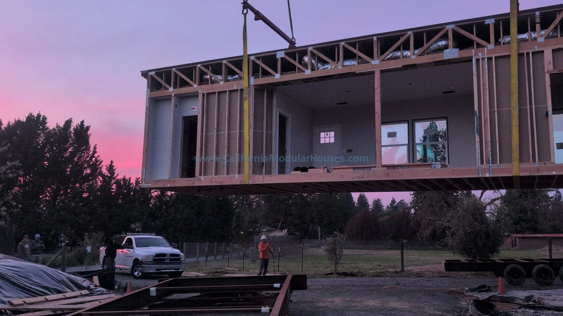 A house under construction, with a wooden frame and walls partially built, is elevated above the ground. A crane is lifting the house into place at sunset, with a pink and purple sky. Several construction workers and a pickup truck are visible on the