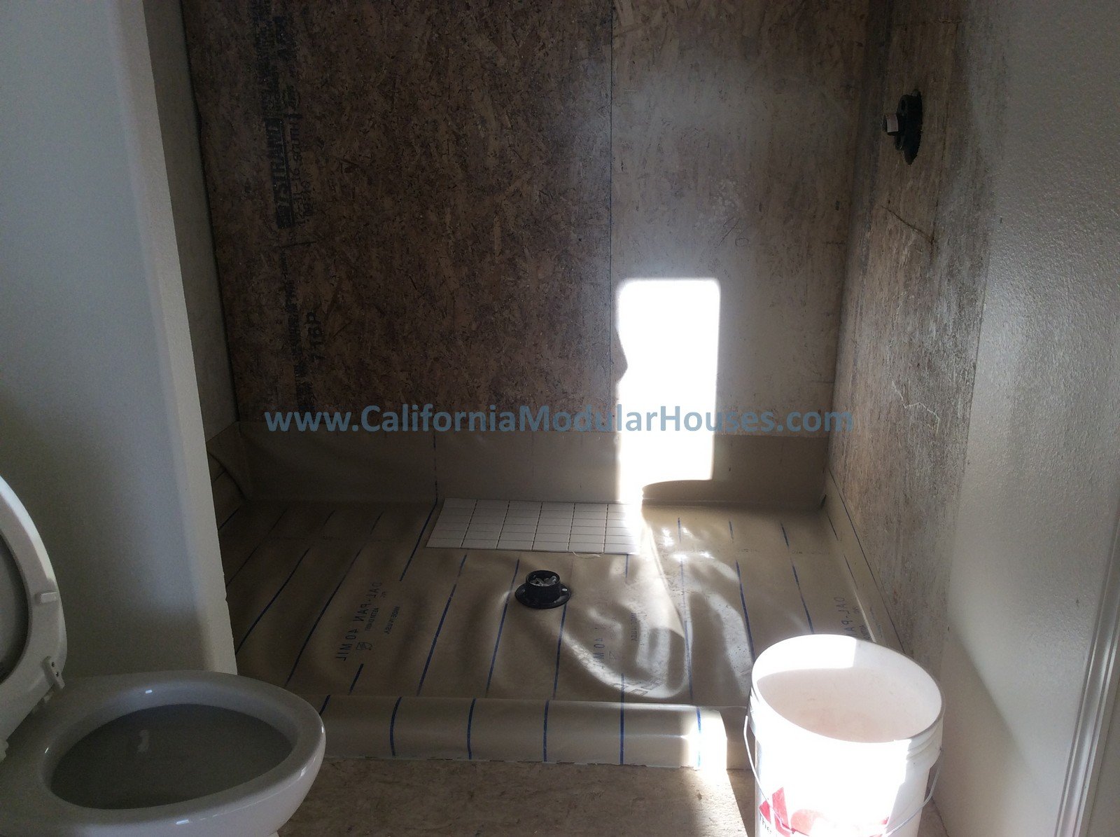 View of a partially renovated bathroom with an empty shower area, a white toilet, a bucket, and construction materials, with unfinished walls and sunlight coming through a small window.