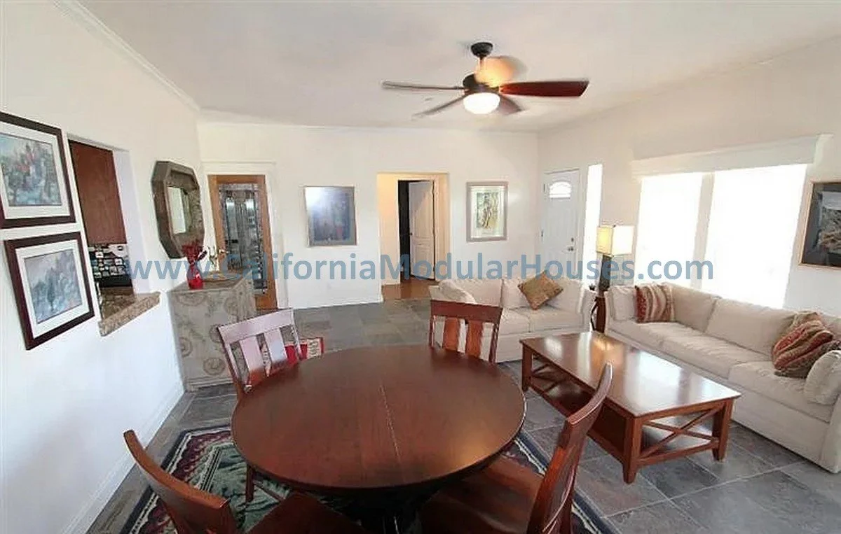 Living room with white walls, a ceiling fan, a round wooden dining table with four chairs, a beige sofa with pillows, a coffee table, and large windows with blinds.