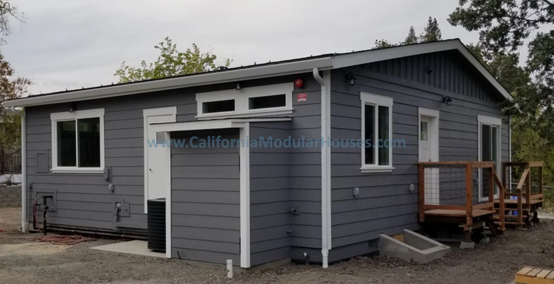 A gray modern tiny house with a small wooden deck and multiple windows, situated on a gravel lot with trees in the background.  Prefab Modular ADU CA
Accessory Dwelling Unit.