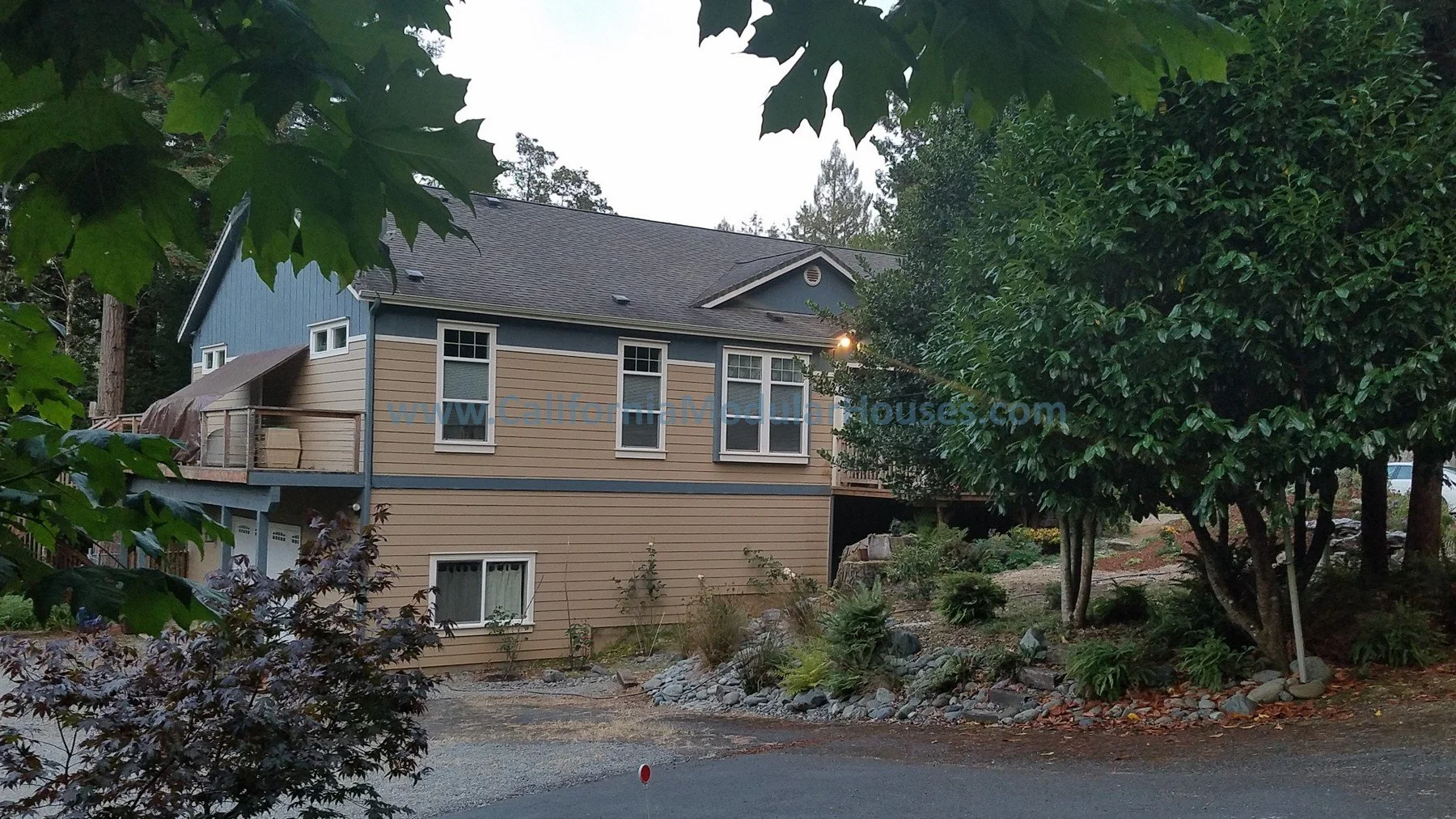 Residential house with tan and blue exterior, multiple windows, trees, and a gravel driveway in a wooded area.  California Modular Houses, Crescent City Modular Home,Modular Home on a Sloped Property. 