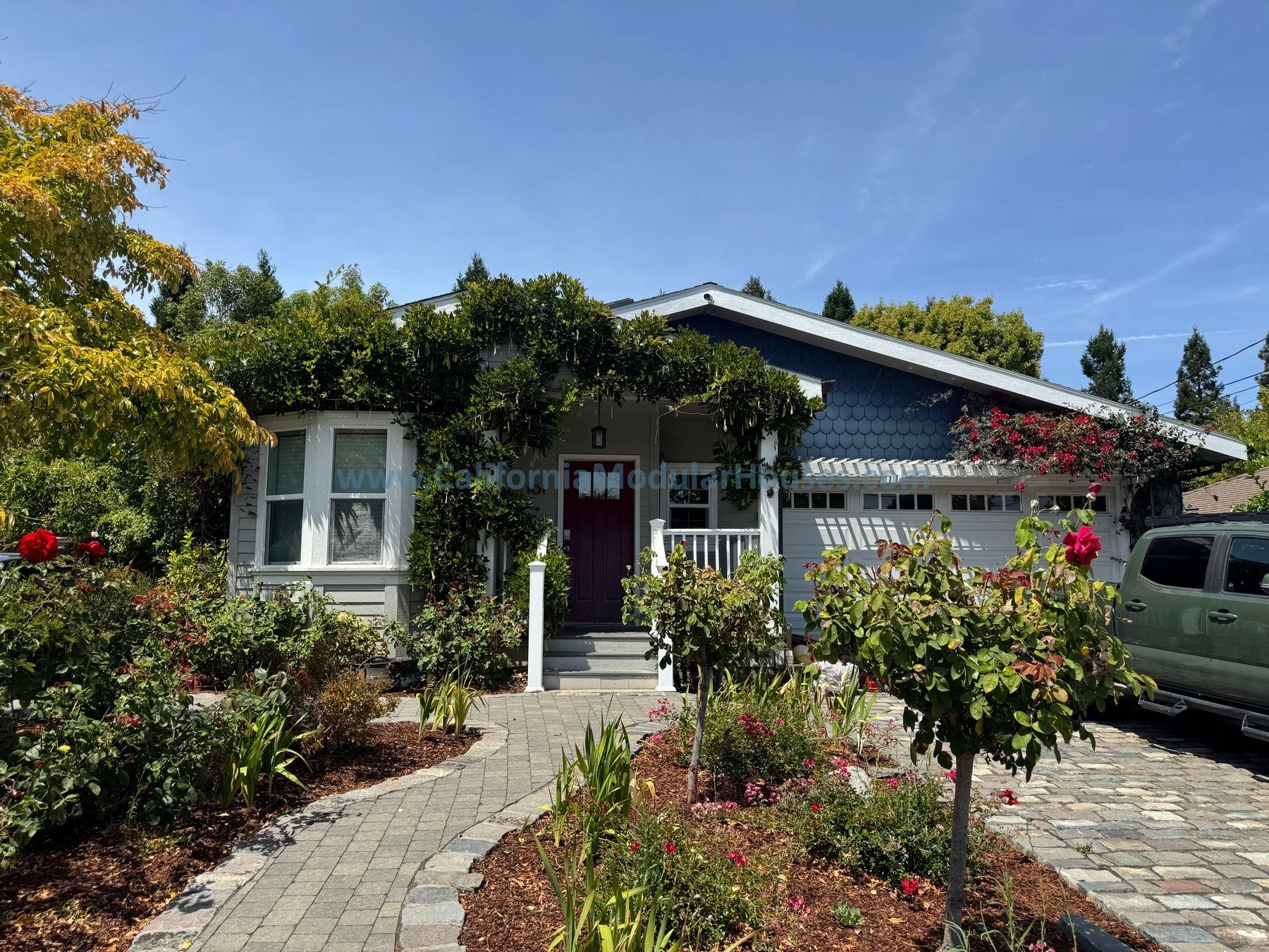 A house with a garden, paved walkway, trees, and flowering plants in front, and a clear blue sky overhead.
