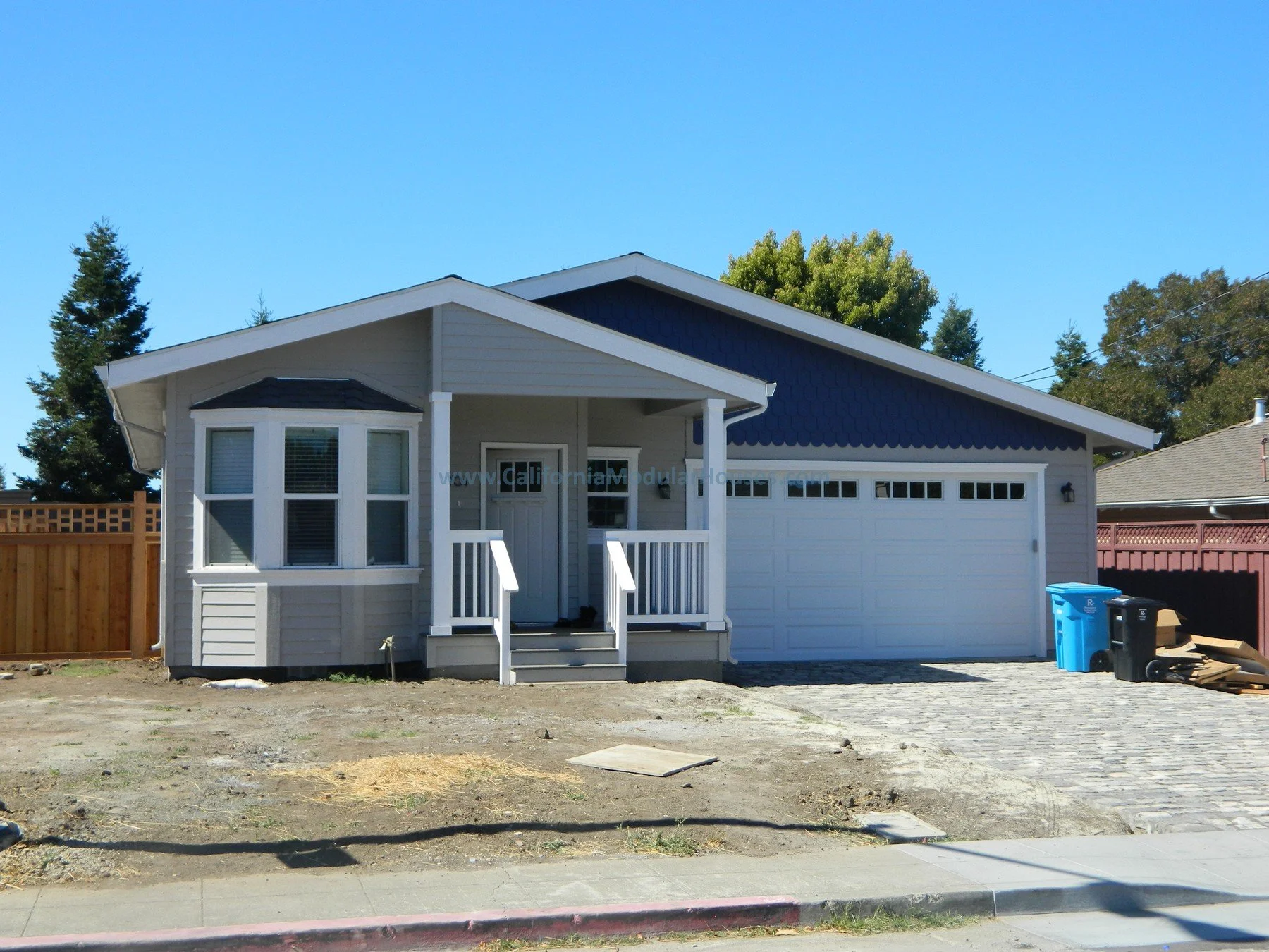 New single-story house with a white garage door, small front porch, and light gray siding, located on a partially paved driveway and lawn area under a clear blue sky.  Modular Homes California, Modular Homes, California Modular Home, 