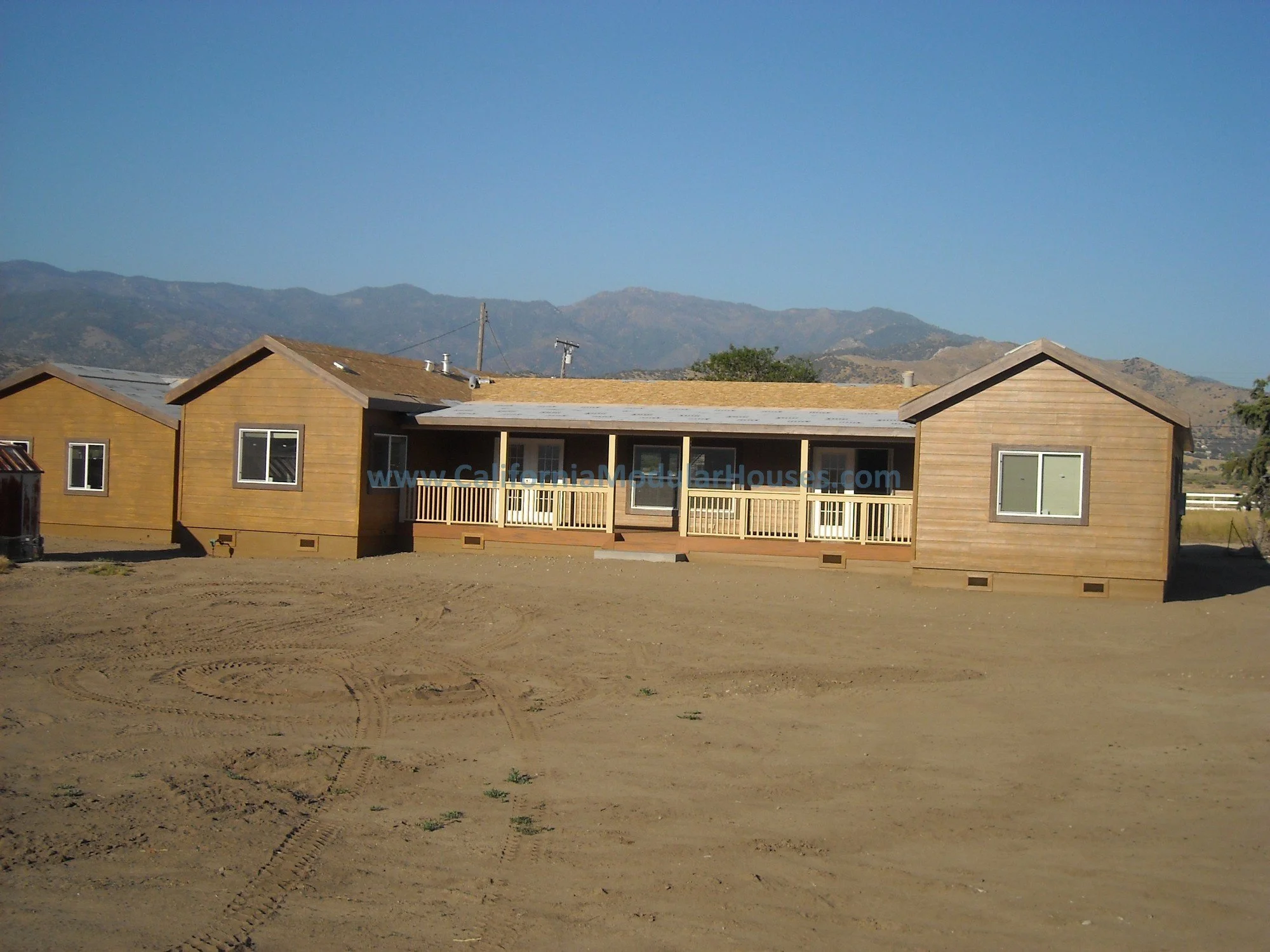 A newly built, single-story wooden house with a small front porch, set on a dirt lot with mountain range in the background.