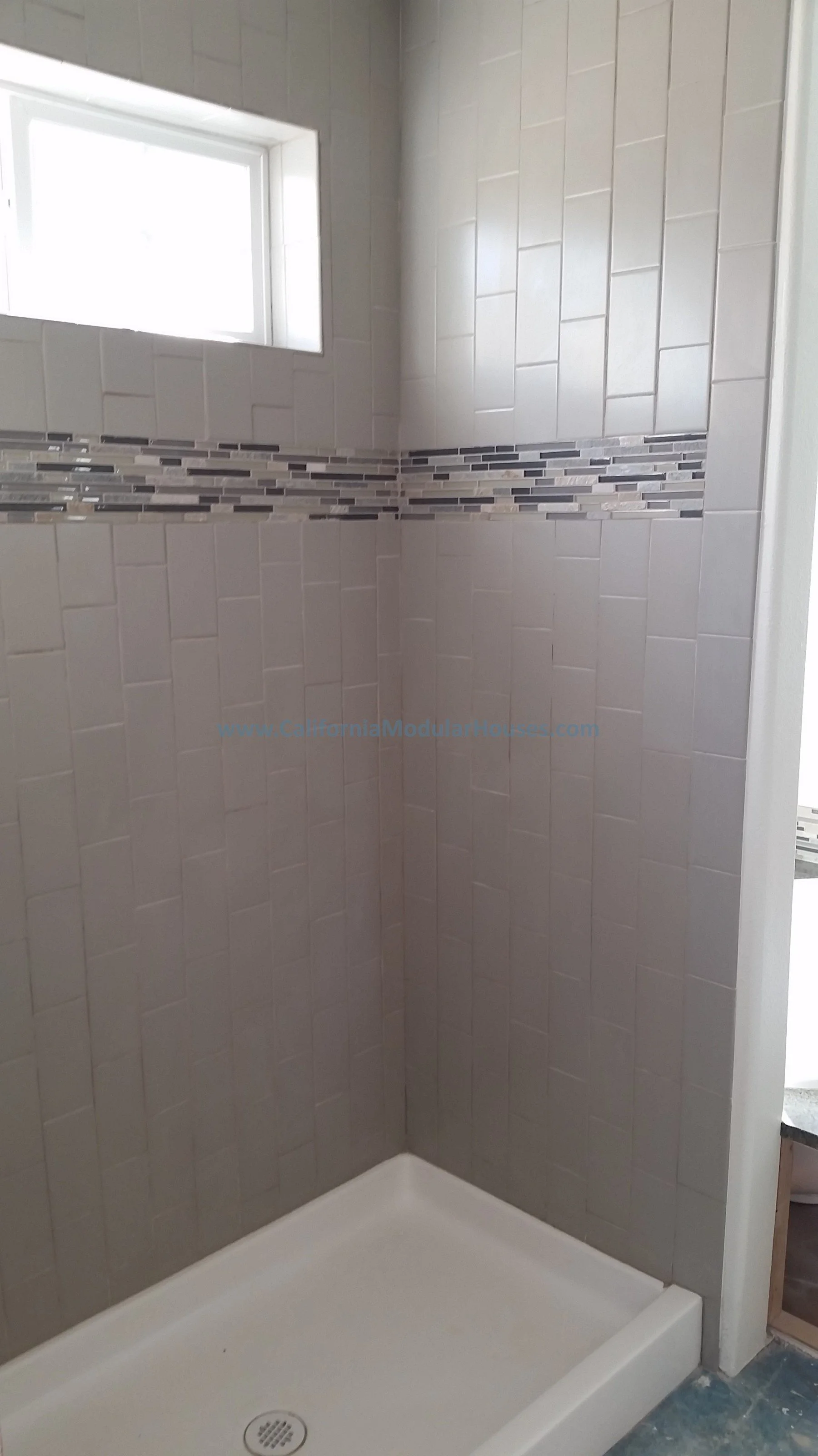 Interior of a shower stall with beige tiles, a decorative horizontal tile strip, and a small window at the top. San Mateo City, San Mateo County, CA. California Modular,