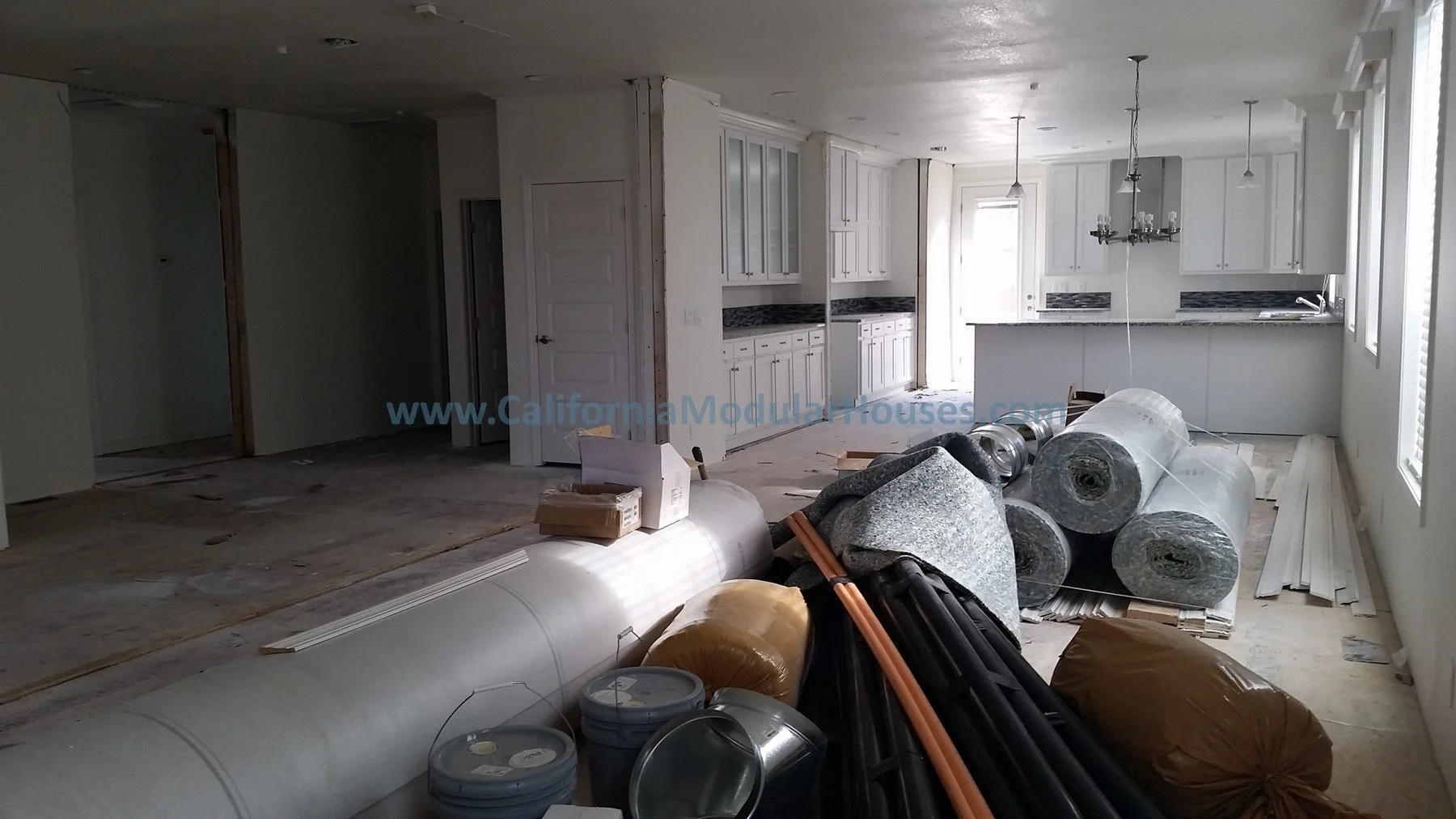 Interior view of a house under construction with white kitchen cabinets and countertops, construction materials, pipes, and insulation rolls in the foreground. Bay Area Prefab Modular Home.