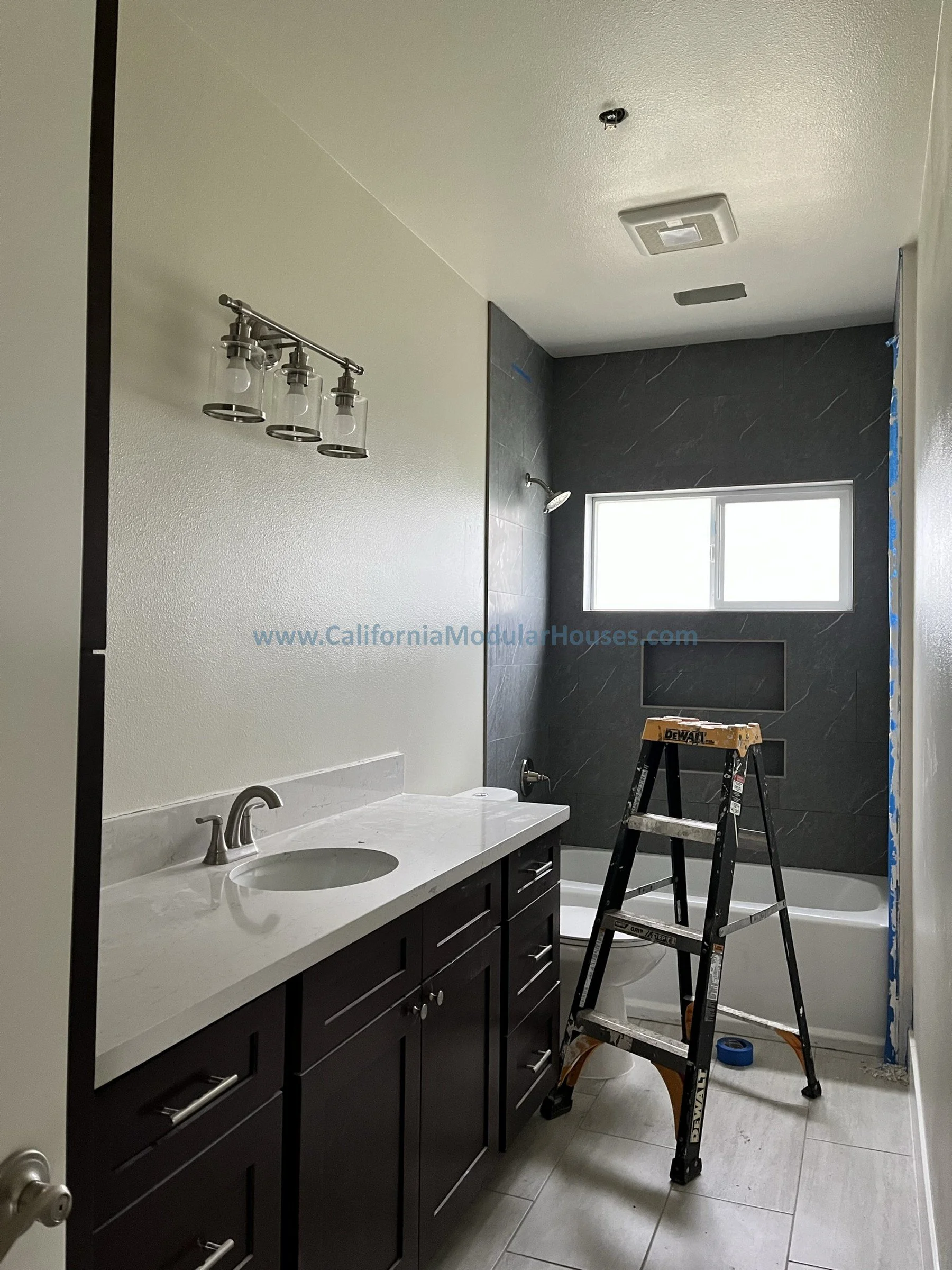 Bathroom under construction with a dark vanity, white countertop, and a shower area with dark gray tiled walls, a window, and a step ladder in the center.