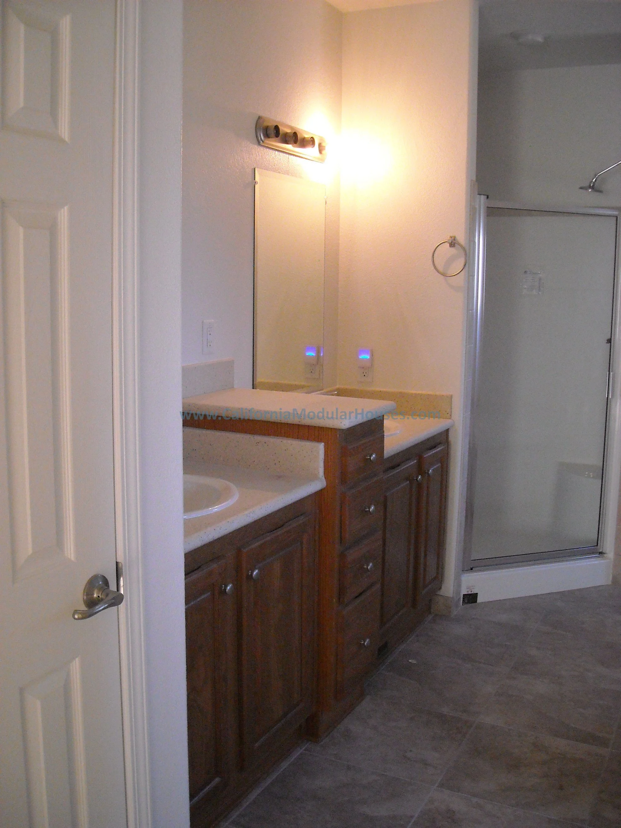 Bathroom vanity with two sinks, a mirror, and a shower stall in the background and a raised area for drawers an a higher cabinet area features of this modular home.  