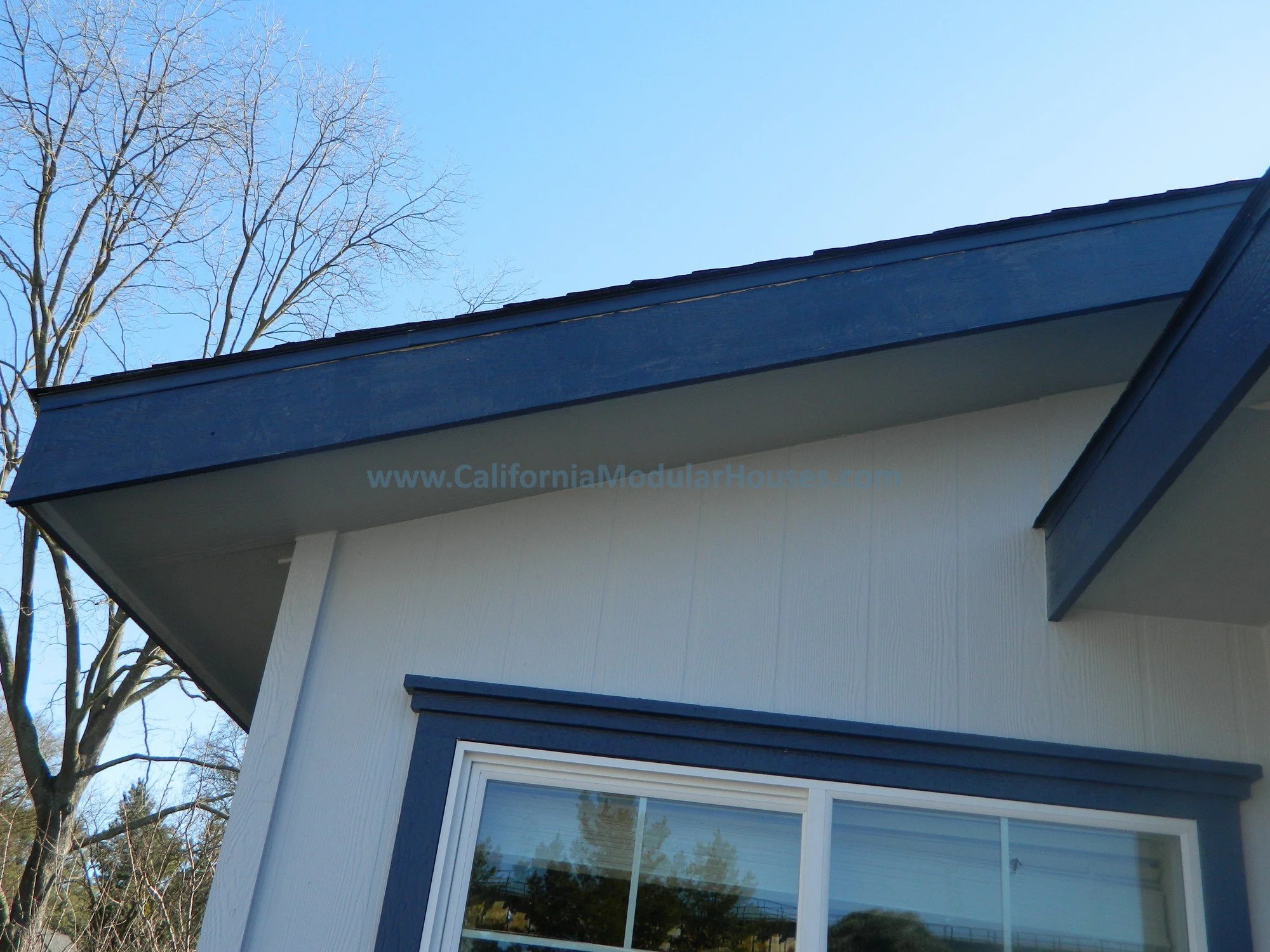 Close-up of a modern house's roof with a blue trim, beige siding, and a window reflecting trees and sky, with leafless trees and clear blue sky in the background.