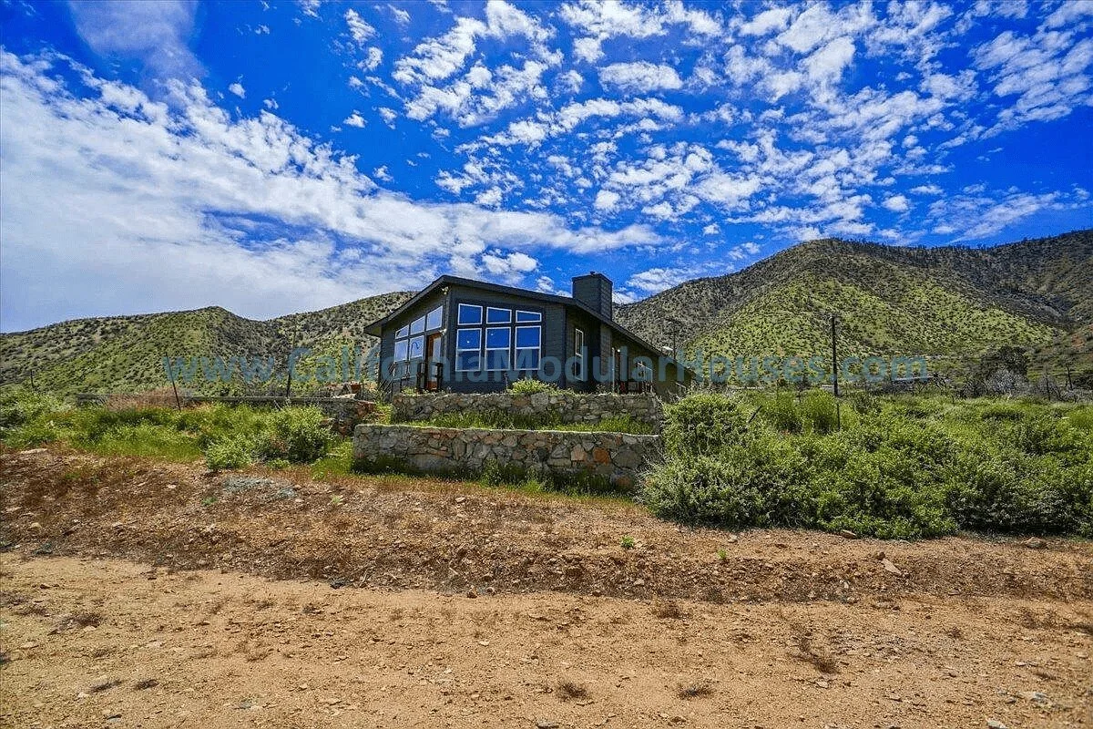 Modern house with large windows on a hill, surrounded by desert and mountains under a partly cloudy sky.