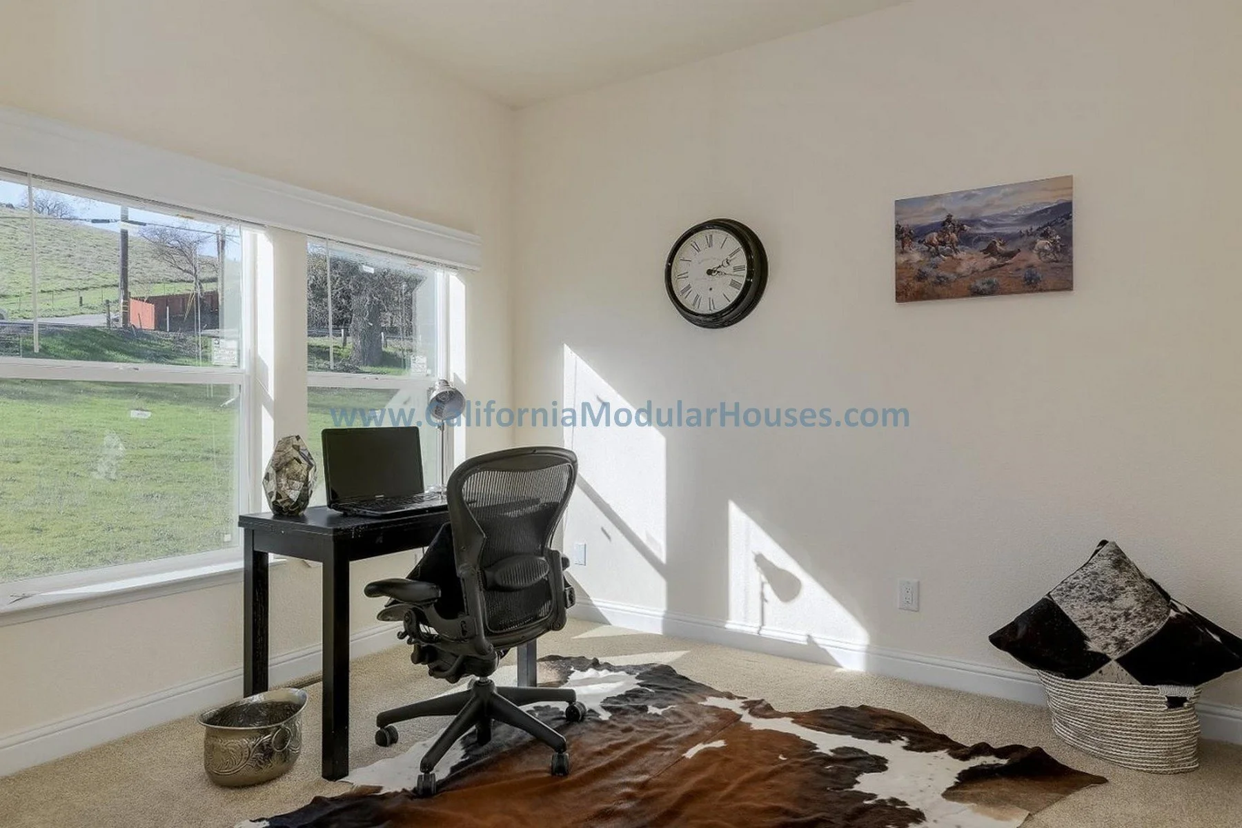 Home office with large window, black desk, ergonomic chair, laptop, decorative items, wall clock, painting, cowhide rug, and a basket with rolled rugs or art pieces.  San Jose, Santa Clara County, CA. Pre-Fabricated Homes, Modular Homes California,