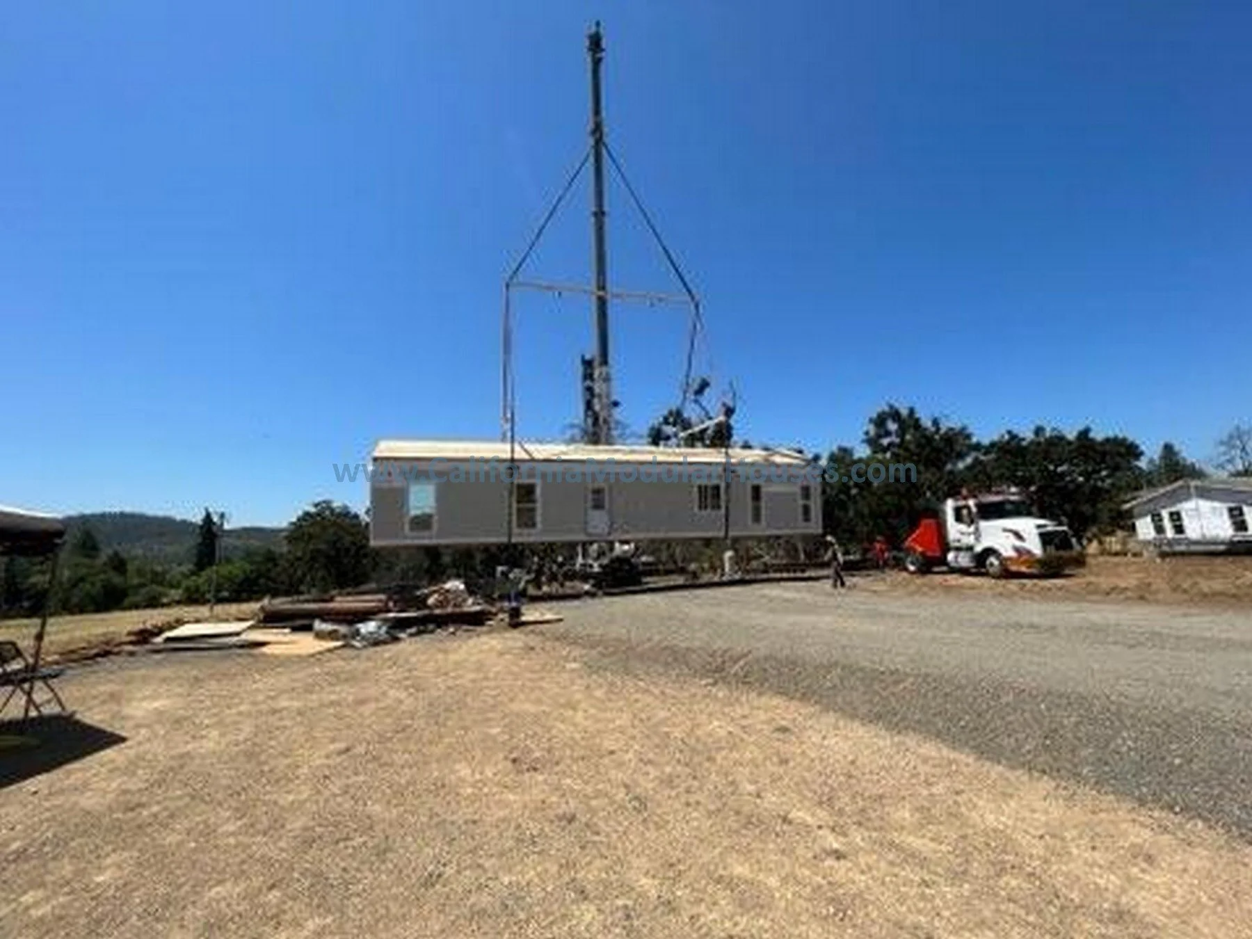 A mobile home is being installed in a rural area with a large antenna mast on top, and construction workers nearby, a semi-truck transporting the home, and other houses in the background under a clear blue sky.
