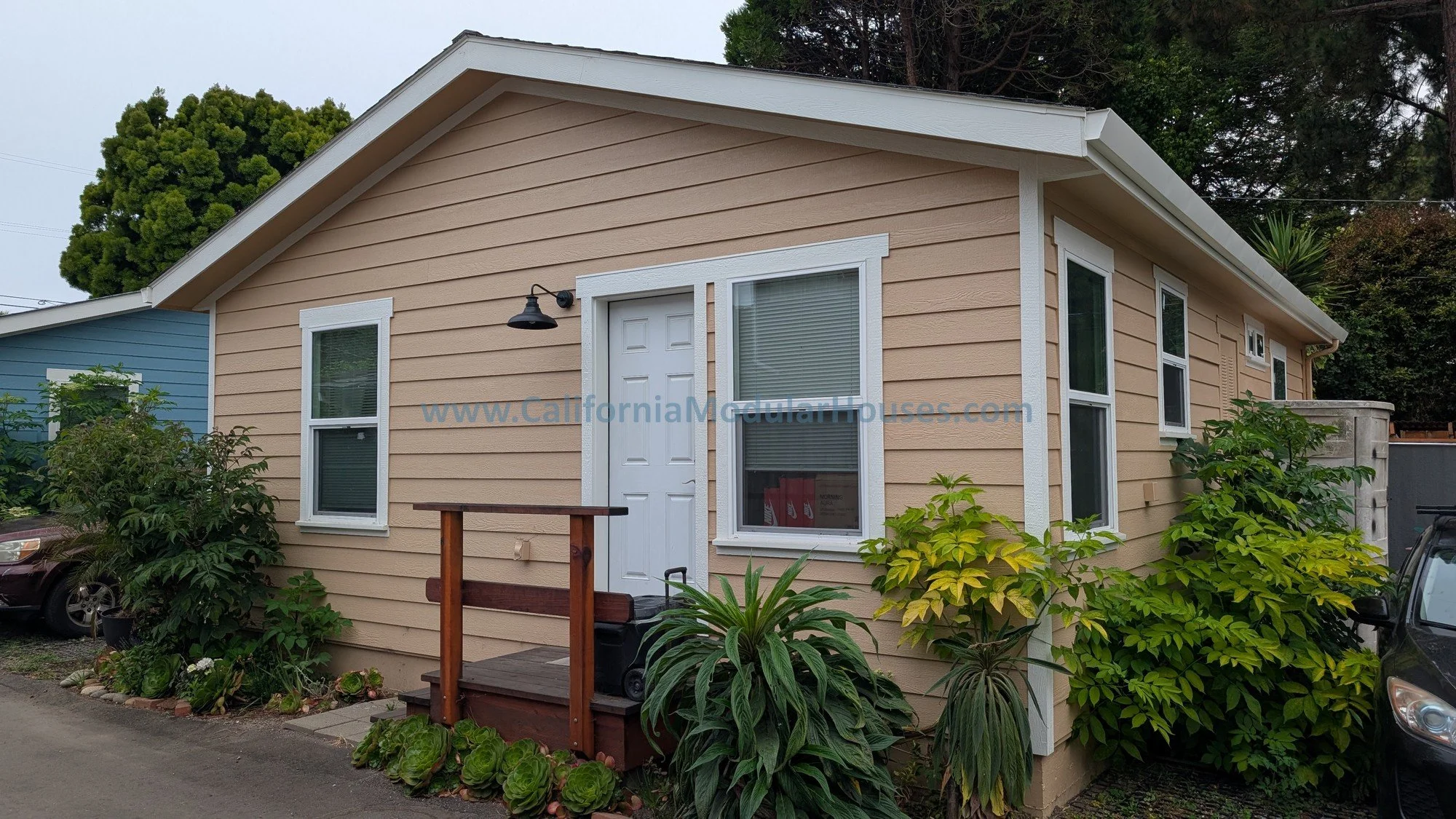 A beige single-story house with white trim and a small front porch with a wooden railing. The house has four windows, with blinds visible through the glass. There are green bushes and plants around the house, and part of a driveway with vehicles on e