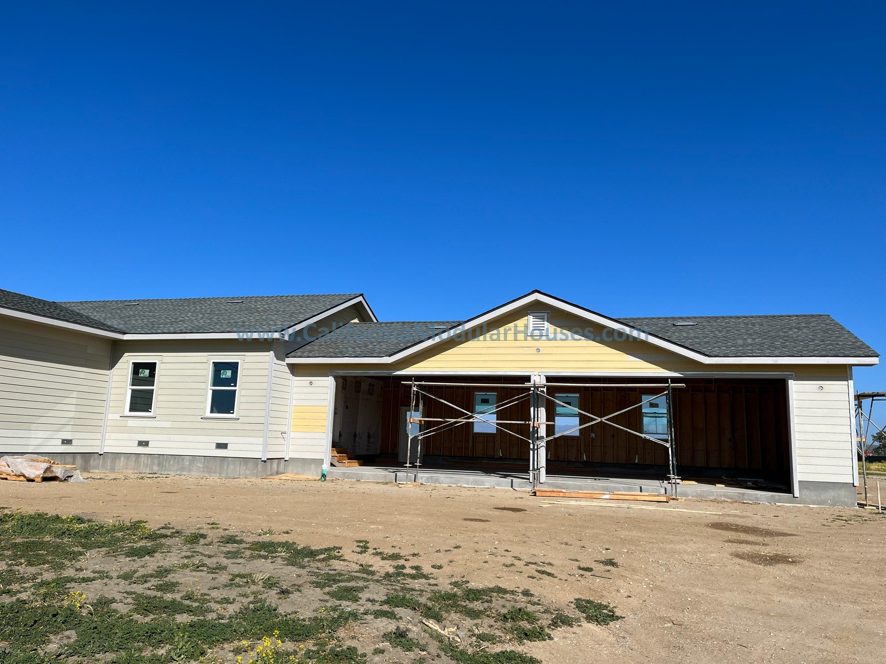 A house under construction with scaffolding in front, white exterior walls, a gray roof, and an open front porch area, against a clear blue sky.