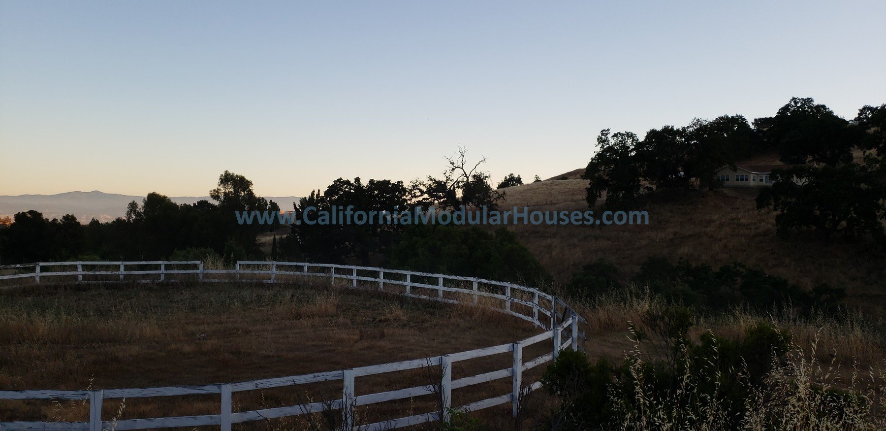 Scenic rural landscape at sunset with rolling hills, scattered trees, a white fence winding through the land, and a house on a hill in the distance, with a clear sky and mountains in the background. Modular Home CA,