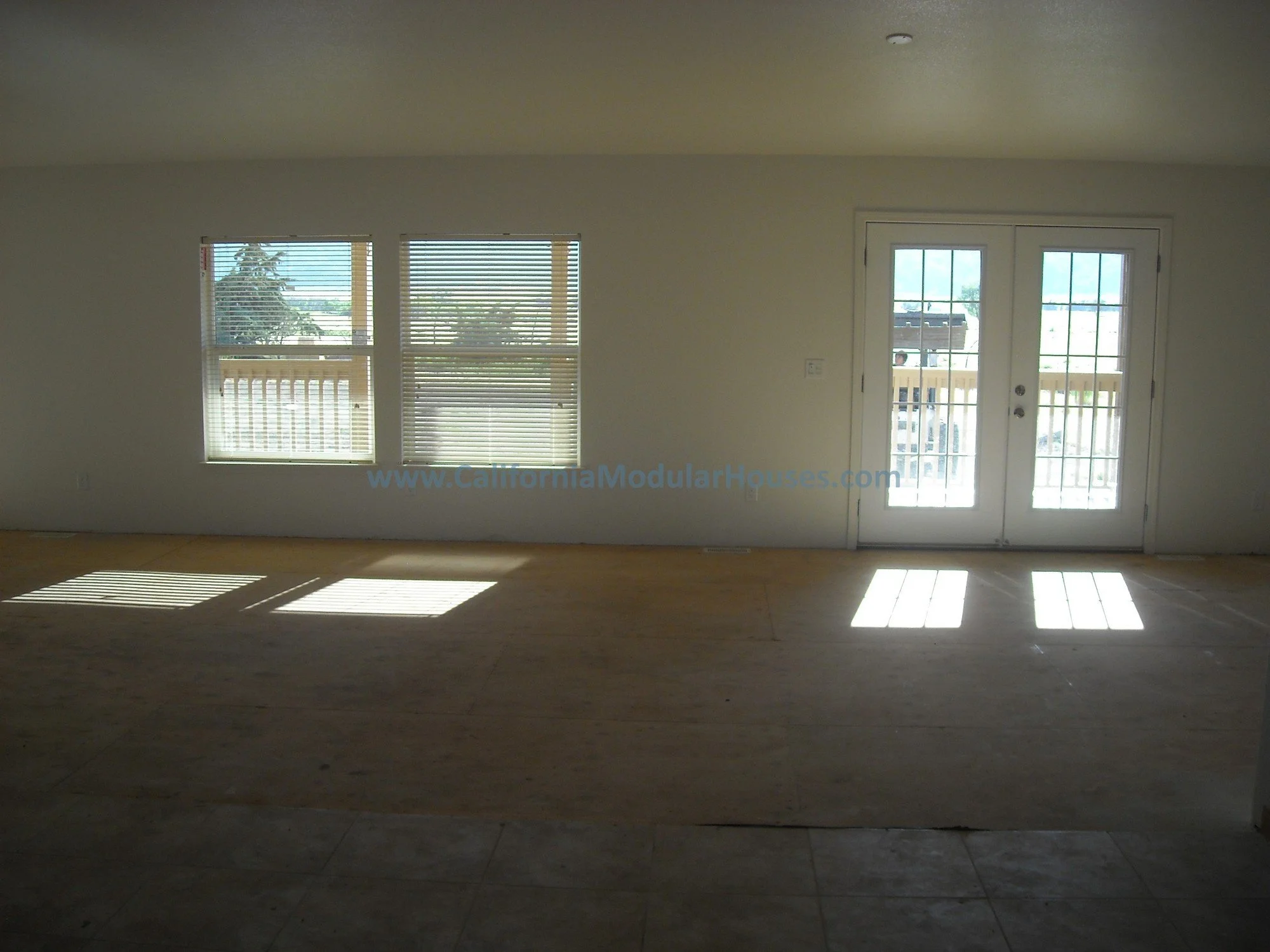 An empty room with two large windows and double glass doors, sunlight casting shadows on the wooden floor, and an outdoor deck visible through the windows and doors.  Look how large this great room is in this modular home! 