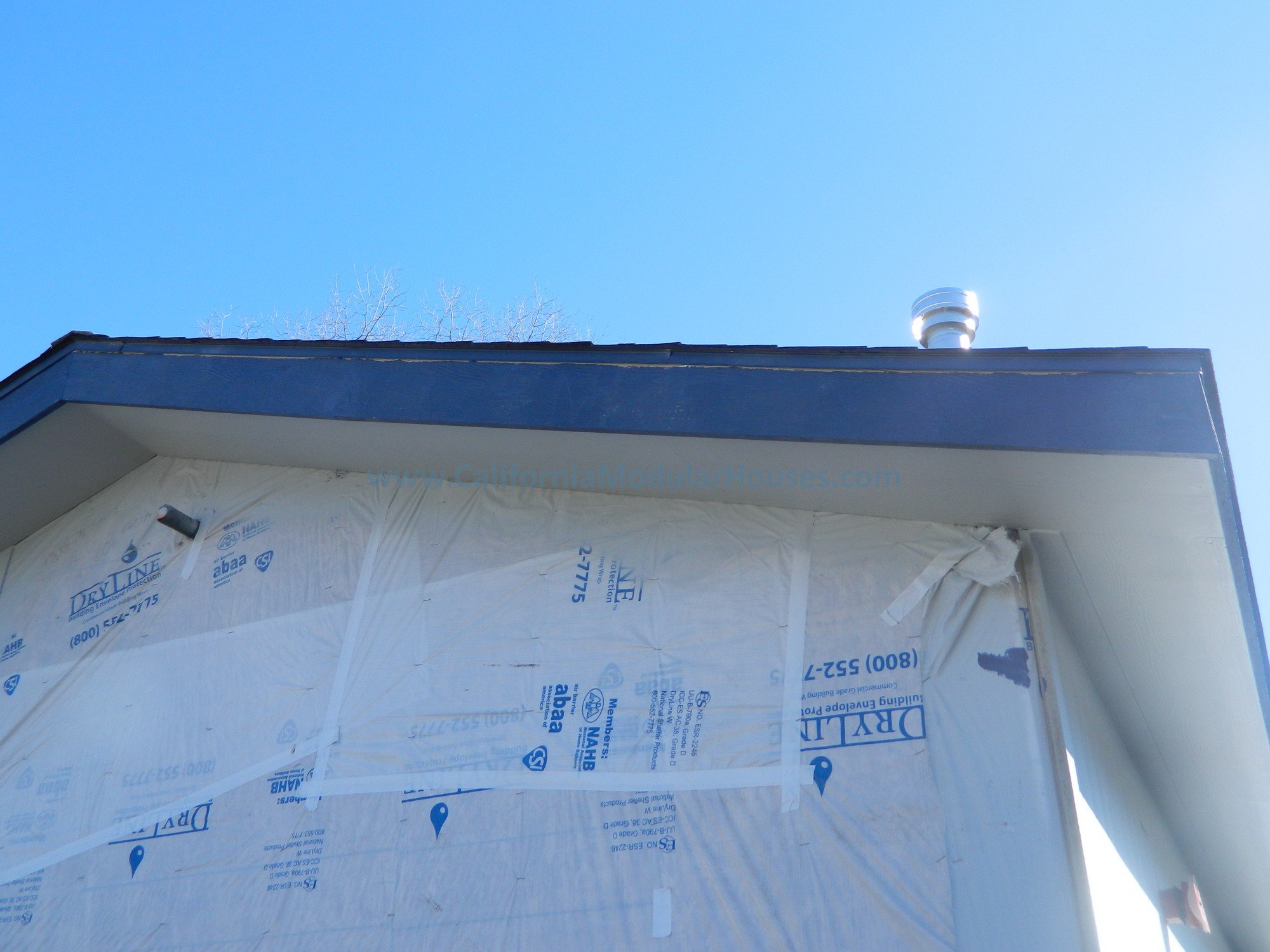 Close-up of the corner of a building under construction, with white weatherproof sheathing, a black roof trim, and a small part of the blue sky visible in the background.
