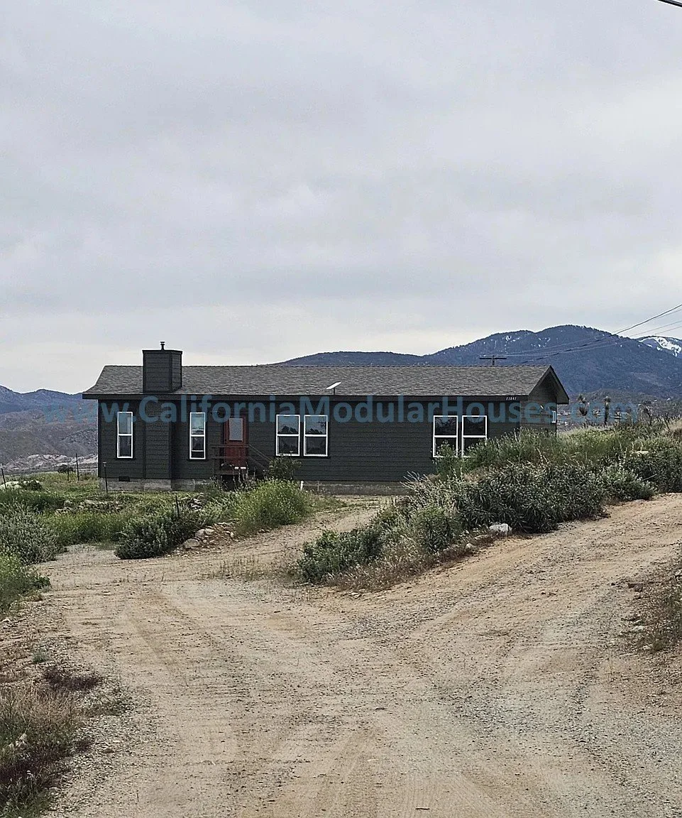 A dark-colored modular house with multiple windows, set in a rural landscape with dirt roads, bushes, and mountains in the background under a cloudy sky.