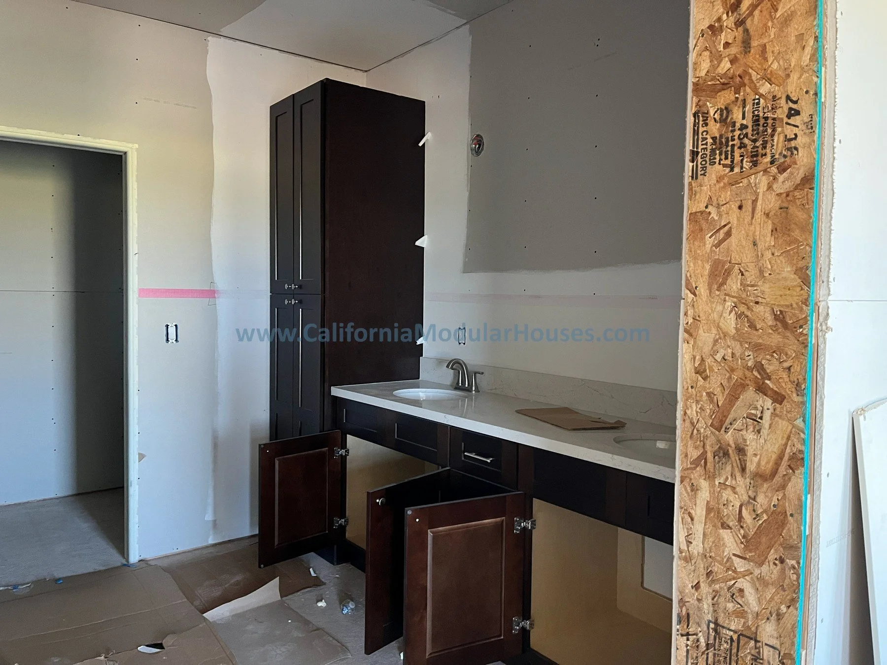 Kitchen under construction with dark wood cabinets, a white countertop, a silver sink, and an unfinished wall with OSB plywood paneling on the right side.