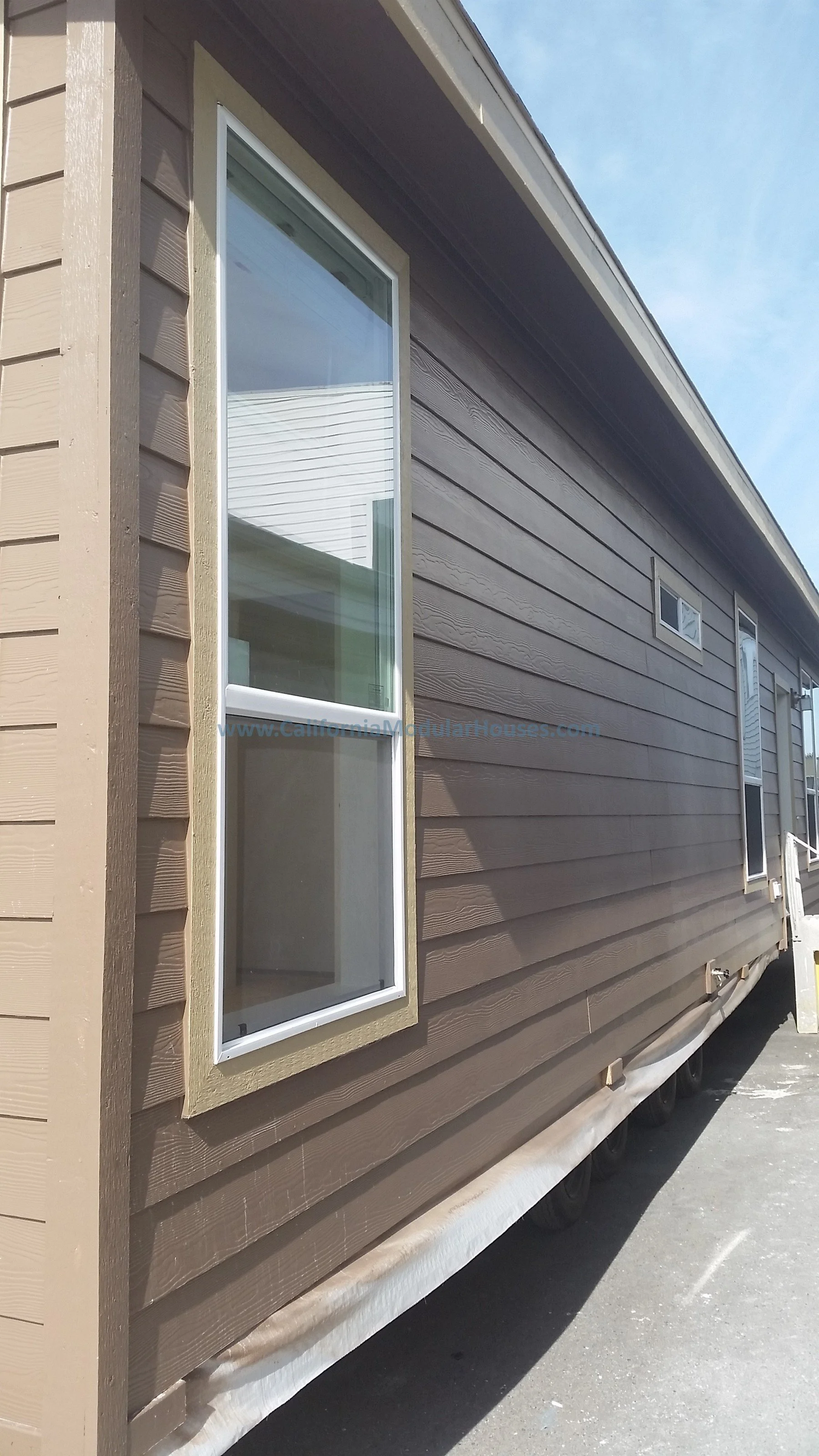 Side view of a newly built modular home for transport with brown siding, a large window, and smaller windows, under a blue sky.  Morgan Hill, Santa Clara County, CA.  Prefab